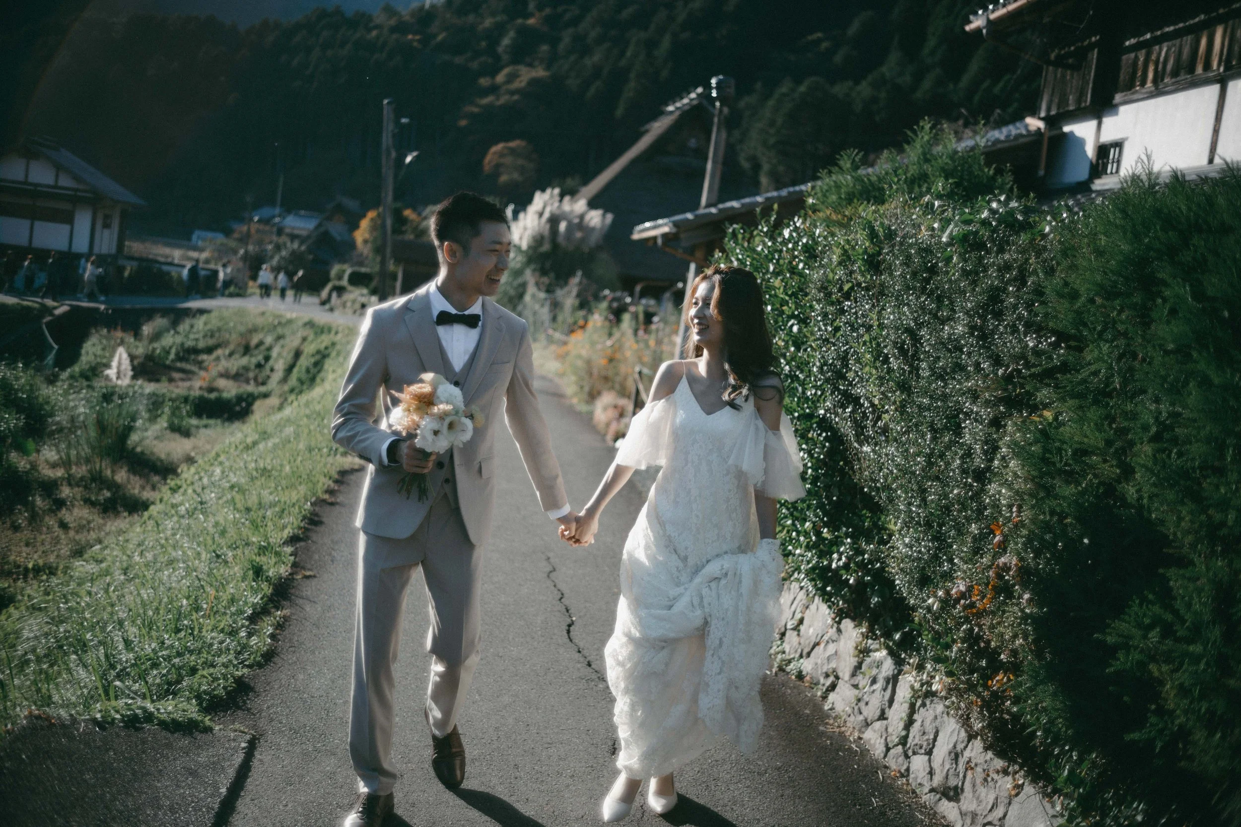 A newlywed couple walking hand in hand along a rural path, with the groom holding a bouquet and wearing a light-colored suit and bow tie, while the bride wears a white lace wedding dress, both smiling at each other. The scene is set against green bus