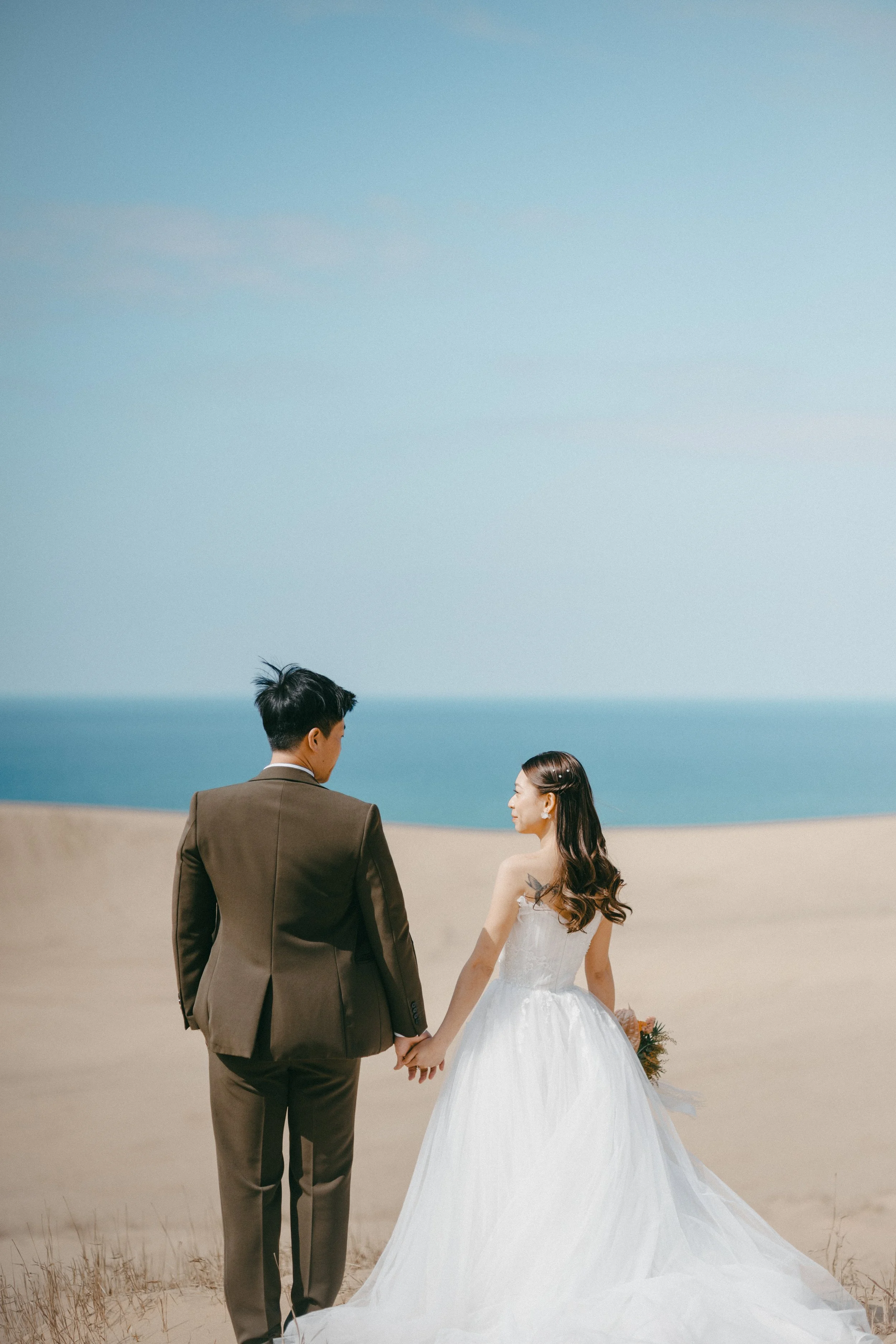 A newlywed couple holding hands on a sandy beach under a clear blue sky, with the ocean in the background.