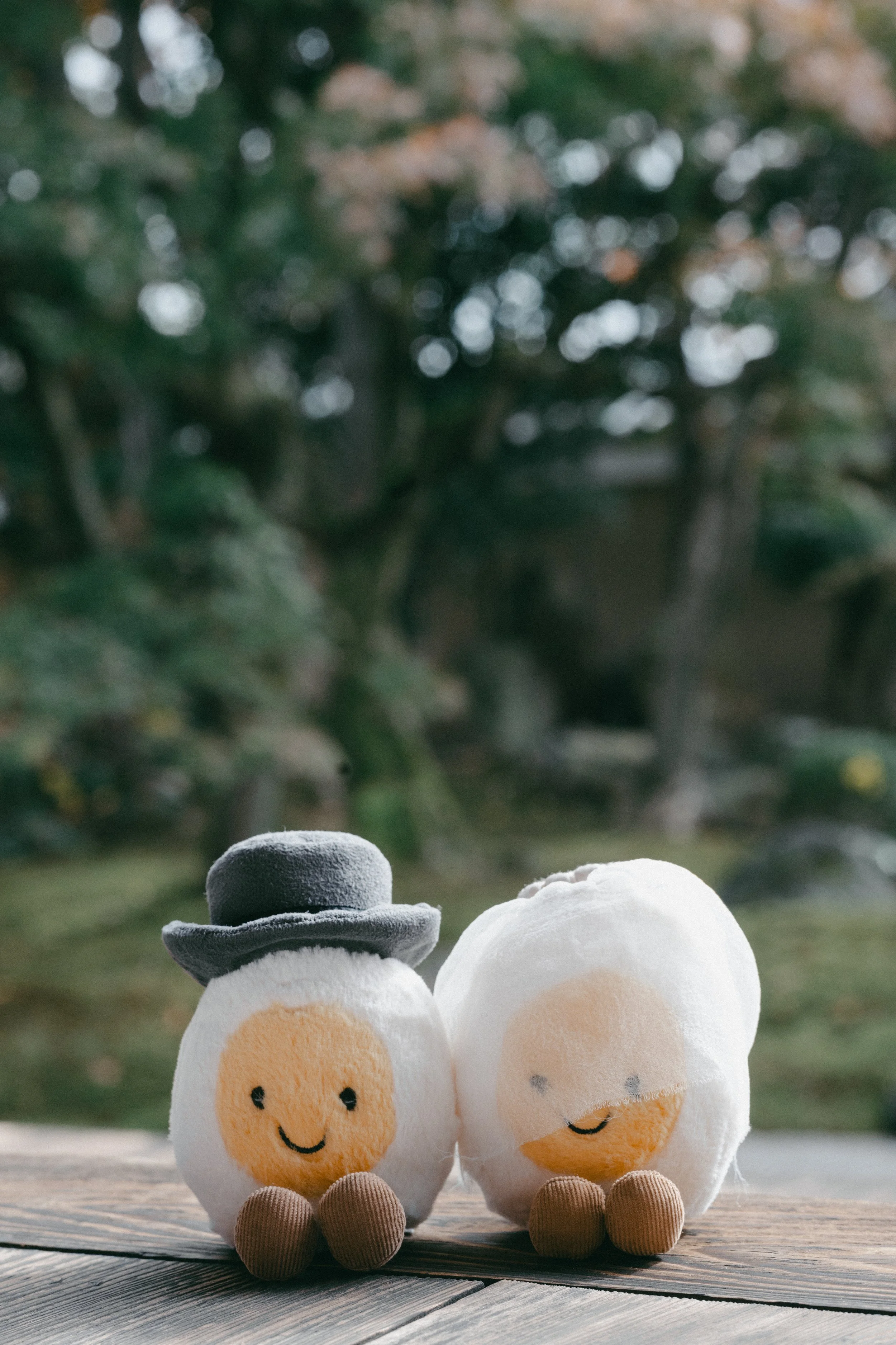 Two plush eggs with smiling faces and small brown feet, one wearing a gray hat, sitting on a wooden surface outdoors with blurred greenery in the background.