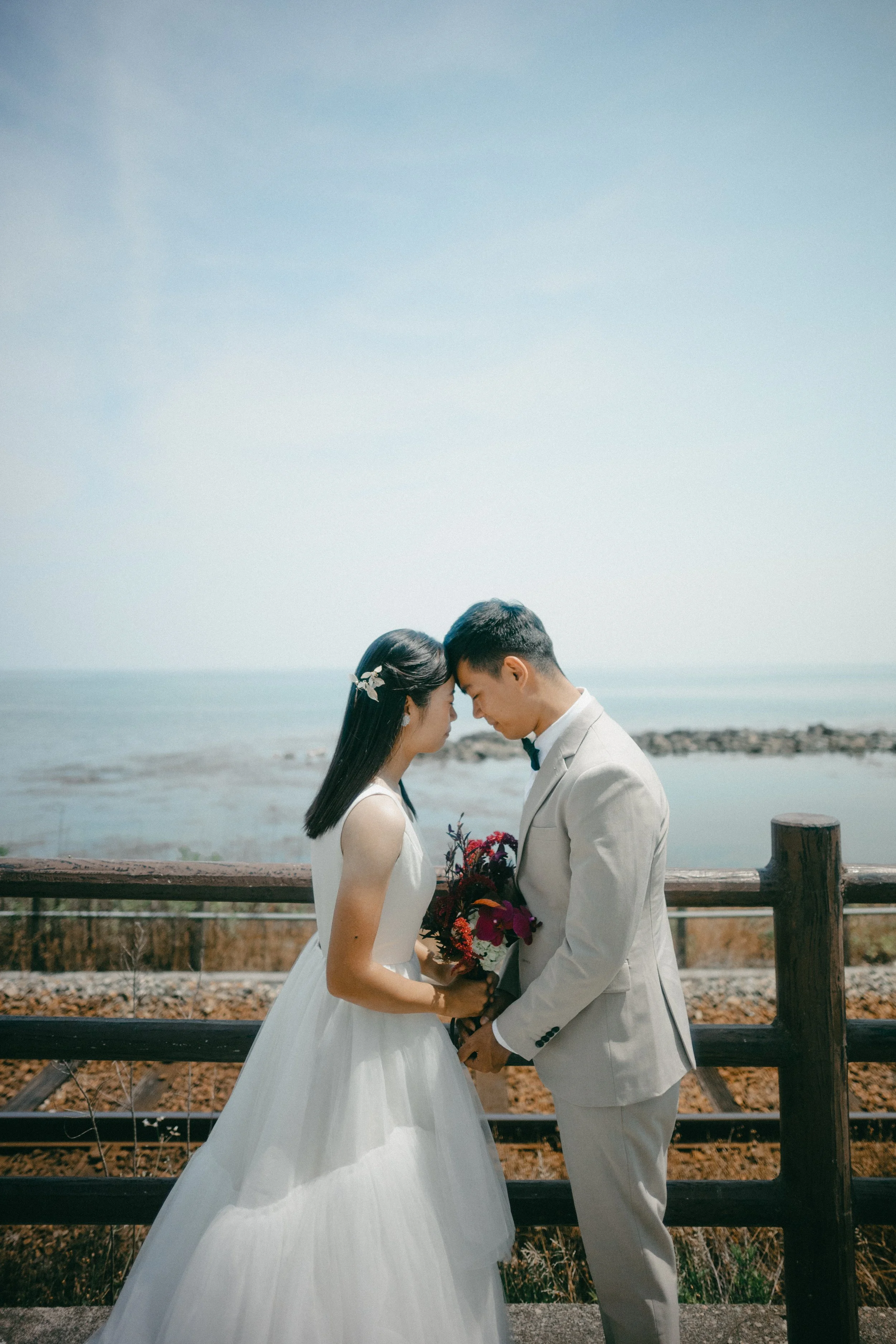 A bride and groom stand close together on a seaside boardwalk, touching foreheads, with the bride holding a bouquet of dark and purple flowers. The scene is bright with a clear sky.