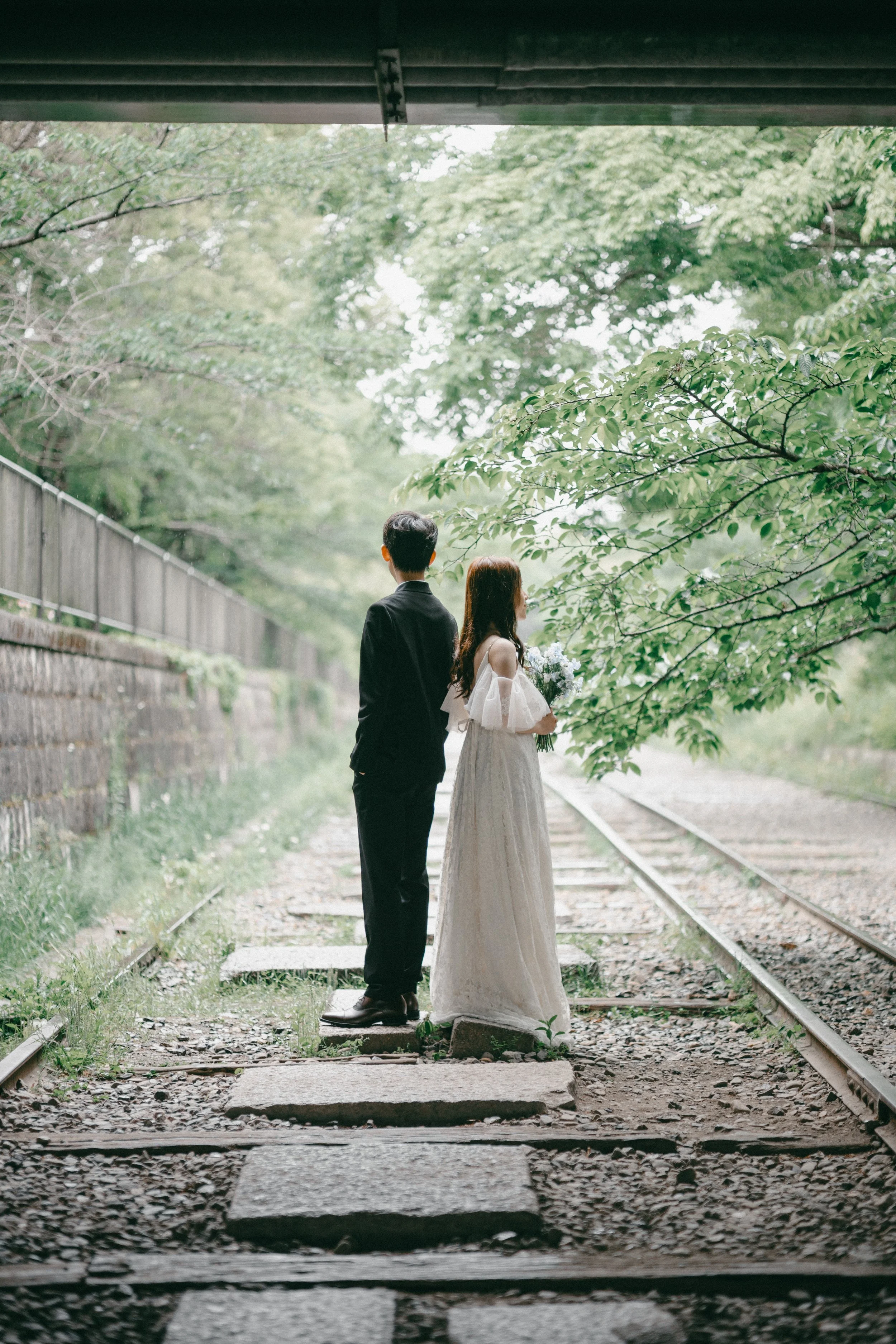 A bride and groom standing together on a small stone platform on railway tracks under a bridge, surrounded by green trees, with the bride holding a bouquet of flowers.