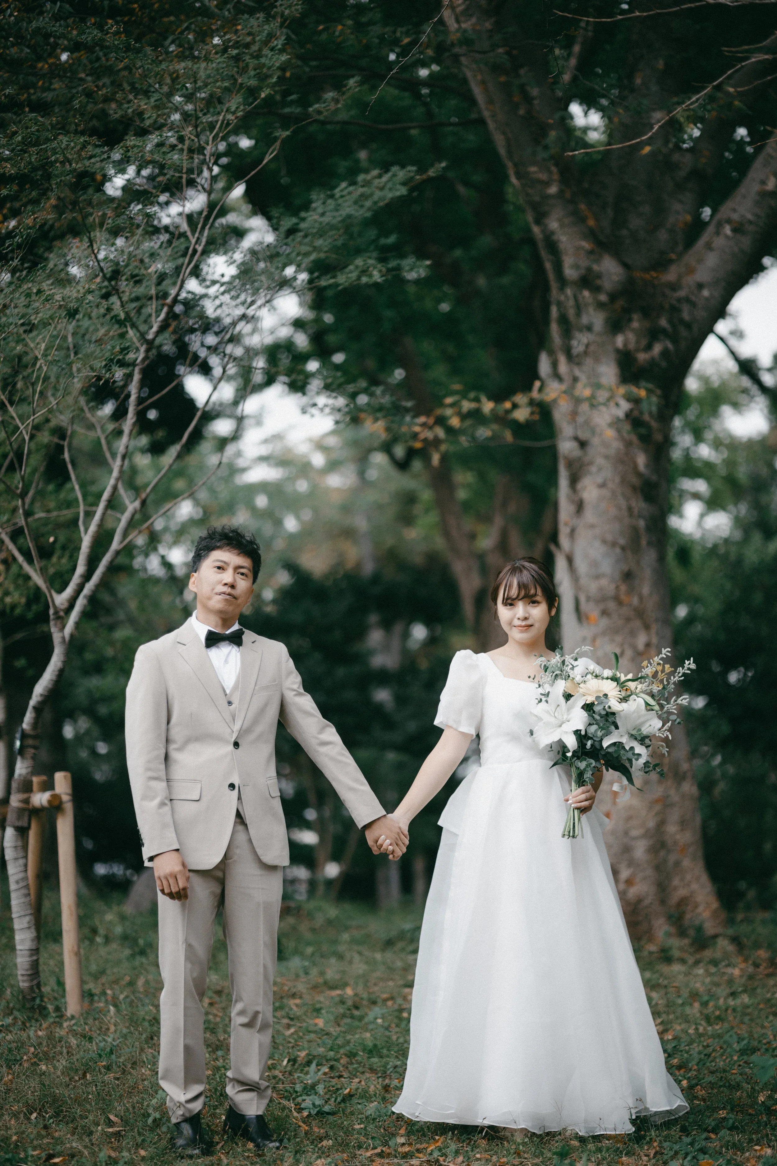 A bride and groom holding hands outdoors in a park, with large trees in the background. The groom is wearing a light beige suit with a bow tie, and the bride is in a white wedding dress holding a bouquet of white flowers.