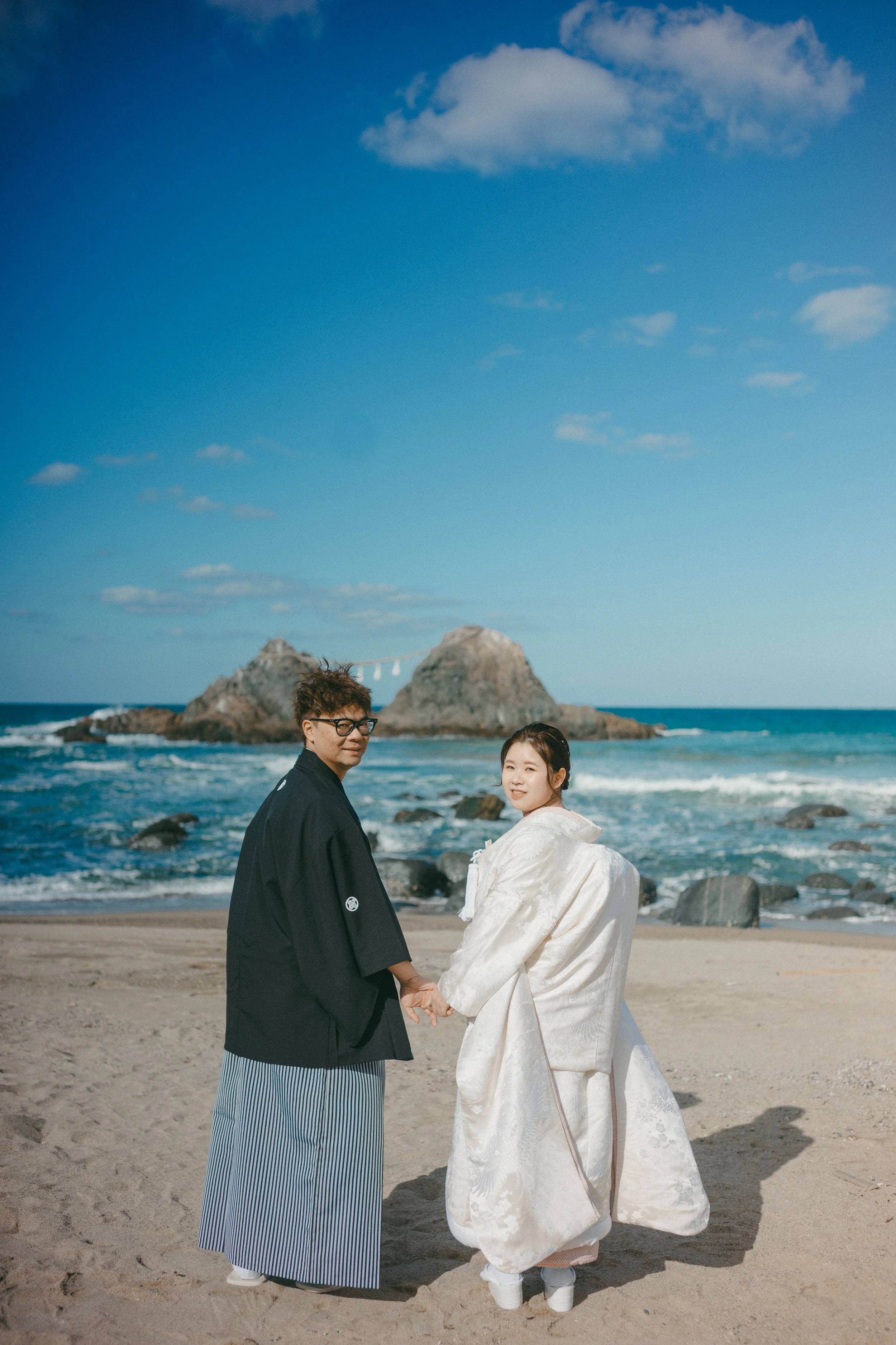 A couple dressed in traditional Japanese wedding attire standing hand in hand on a beach with rocks and ocean in the background under a blue sky.
