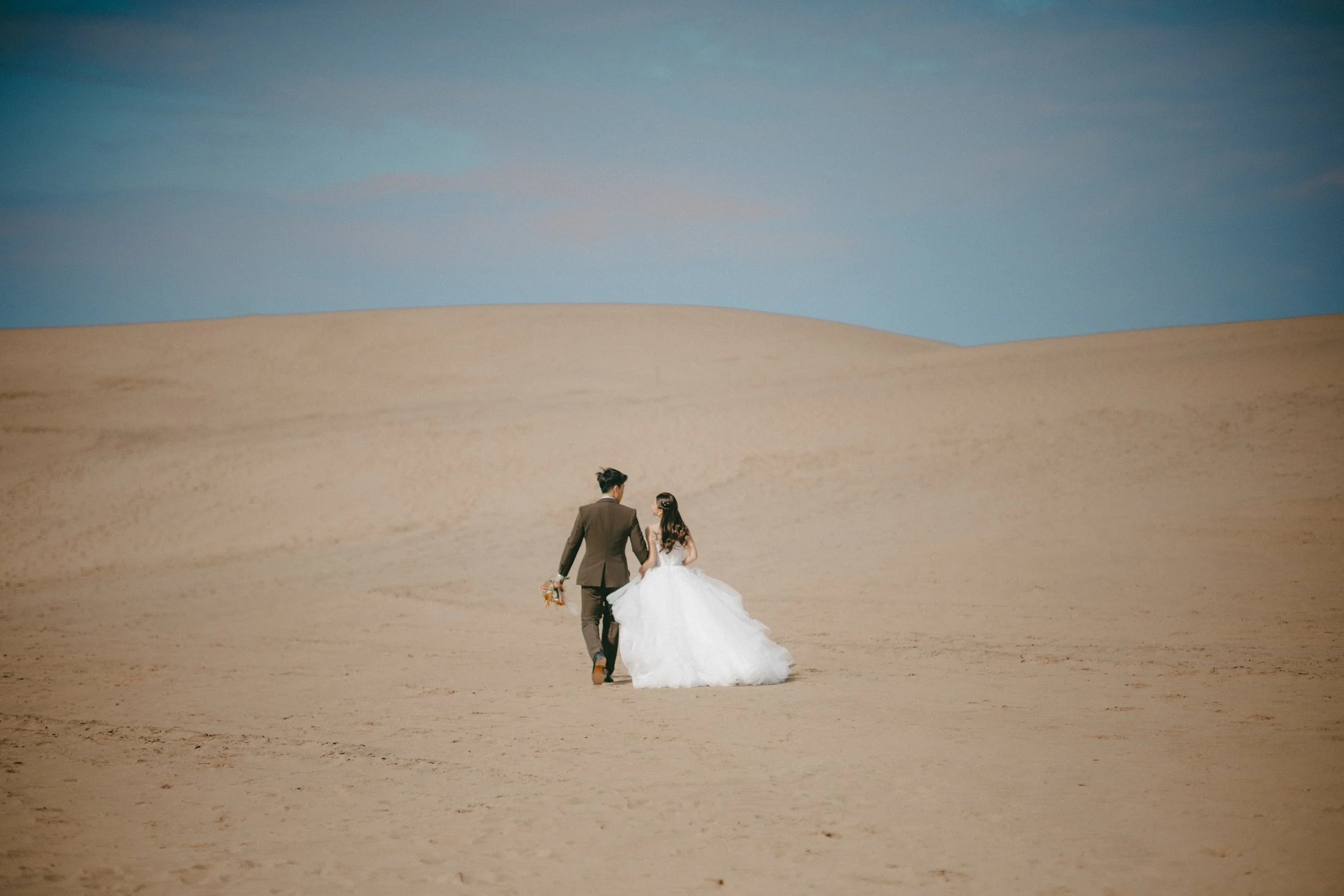 A couple dressed in wedding attire walking hand in hand across a vast sandy desert under a blue sky.