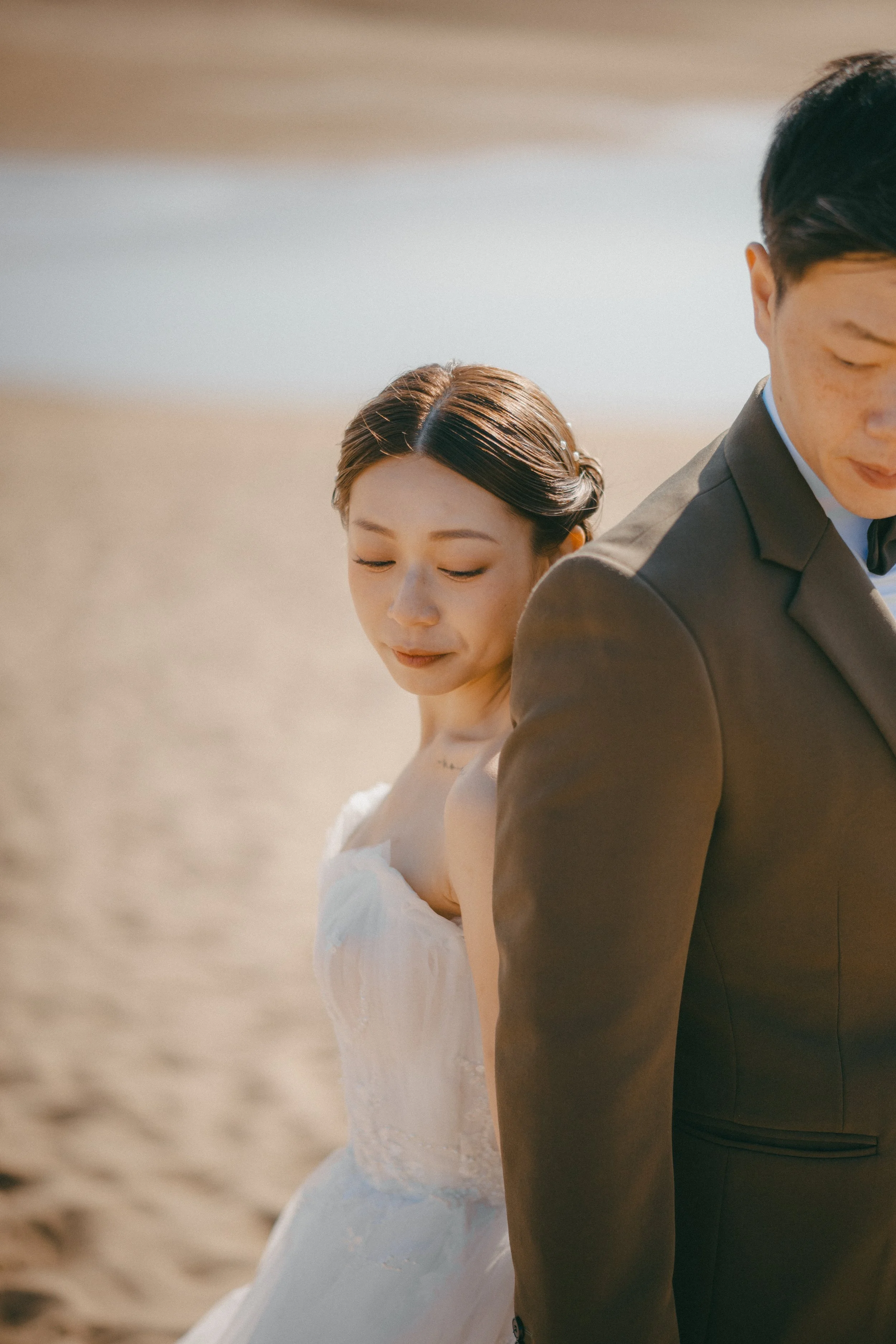 A bride and groom standing back-to-back on a sandy beach with the ocean in the background, both with their eyes closed.