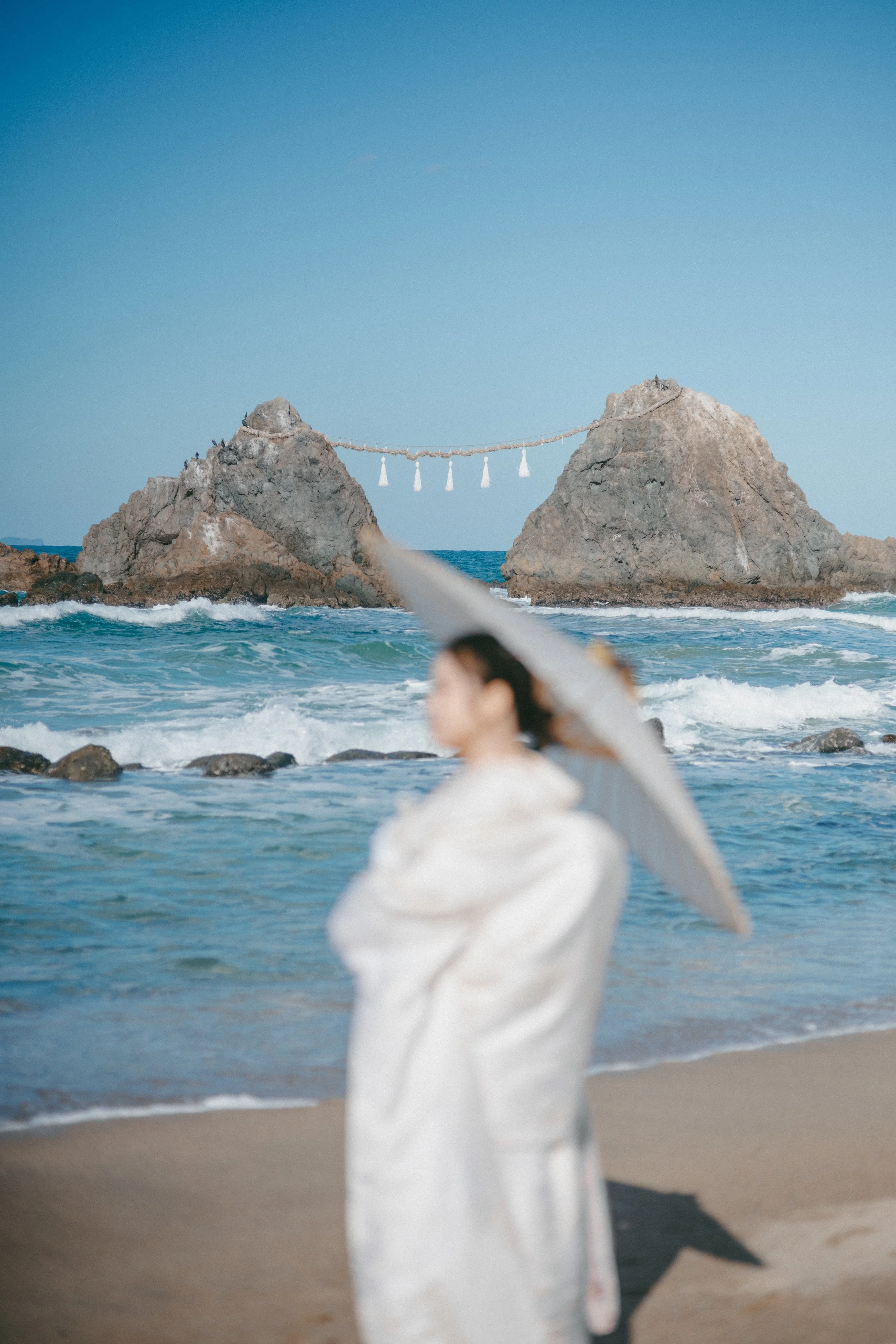 A woman in traditional attire holding a parasol on the beach, with rocky sea stacks and a shimenawa rope in the background.