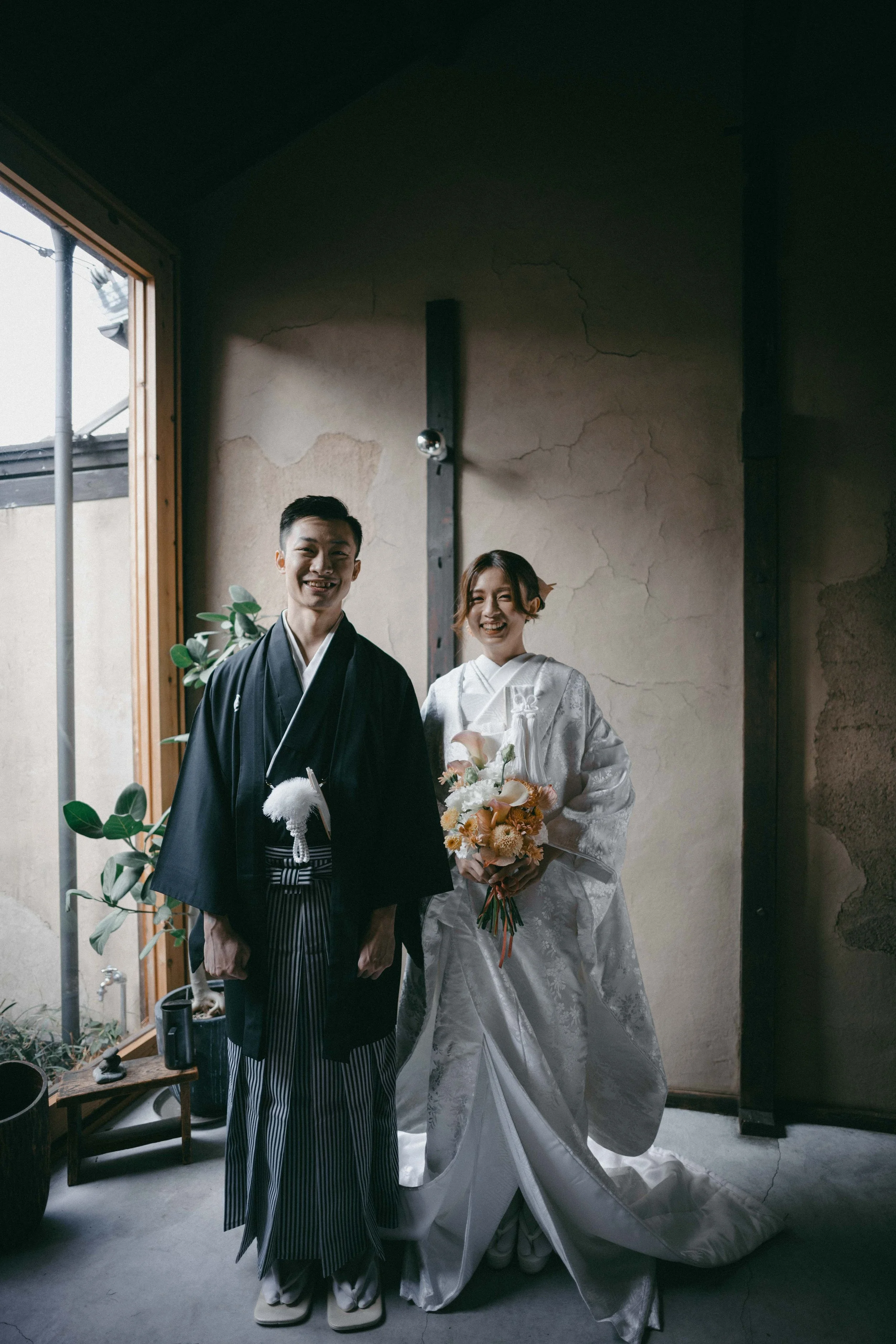 A couple dressed in traditional Japanese wedding attire standing indoors near a window, smiling, with the woman holding a bouquet of flowers.