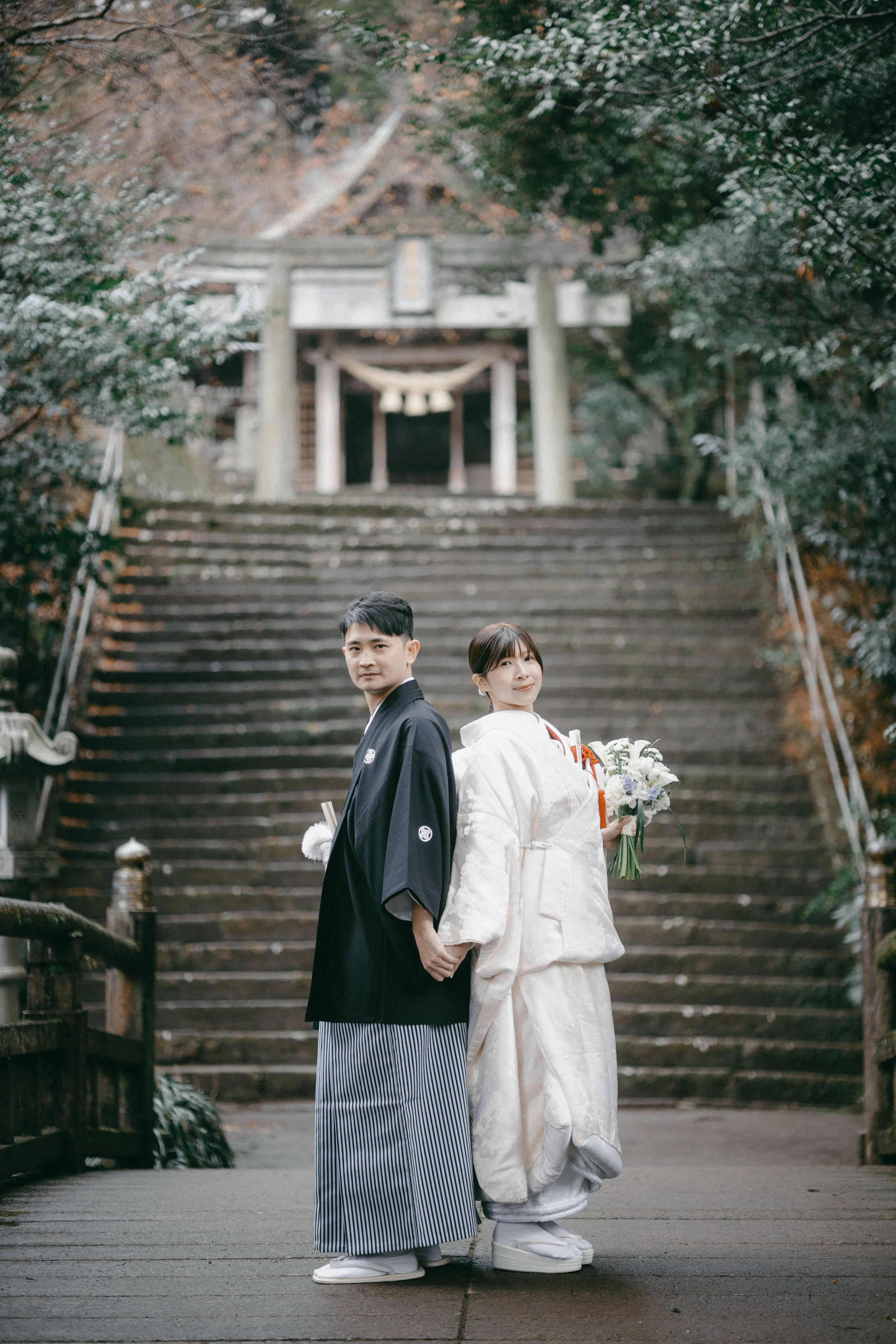 A bride and groom holding hands and standing on a wooden bridge in front of a traditional Japanese shrine with stone steps and a wooden torii gate, dressed in wedding attire.