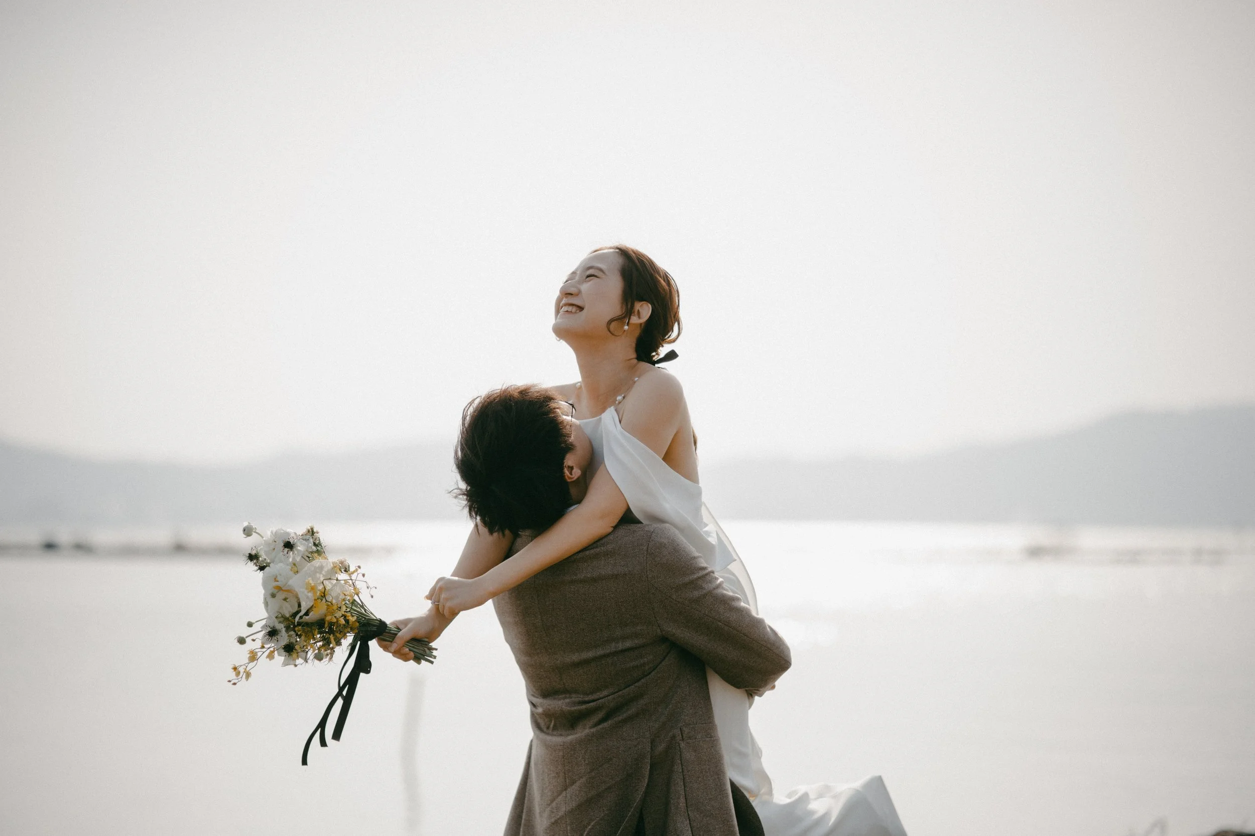 A joyful woman in a white dress is lying over a man dressed in a brown suit, at the beach with a calm sea and mountains in the background. She is holding a bouquet of flowers and smiling with her eyes closed.