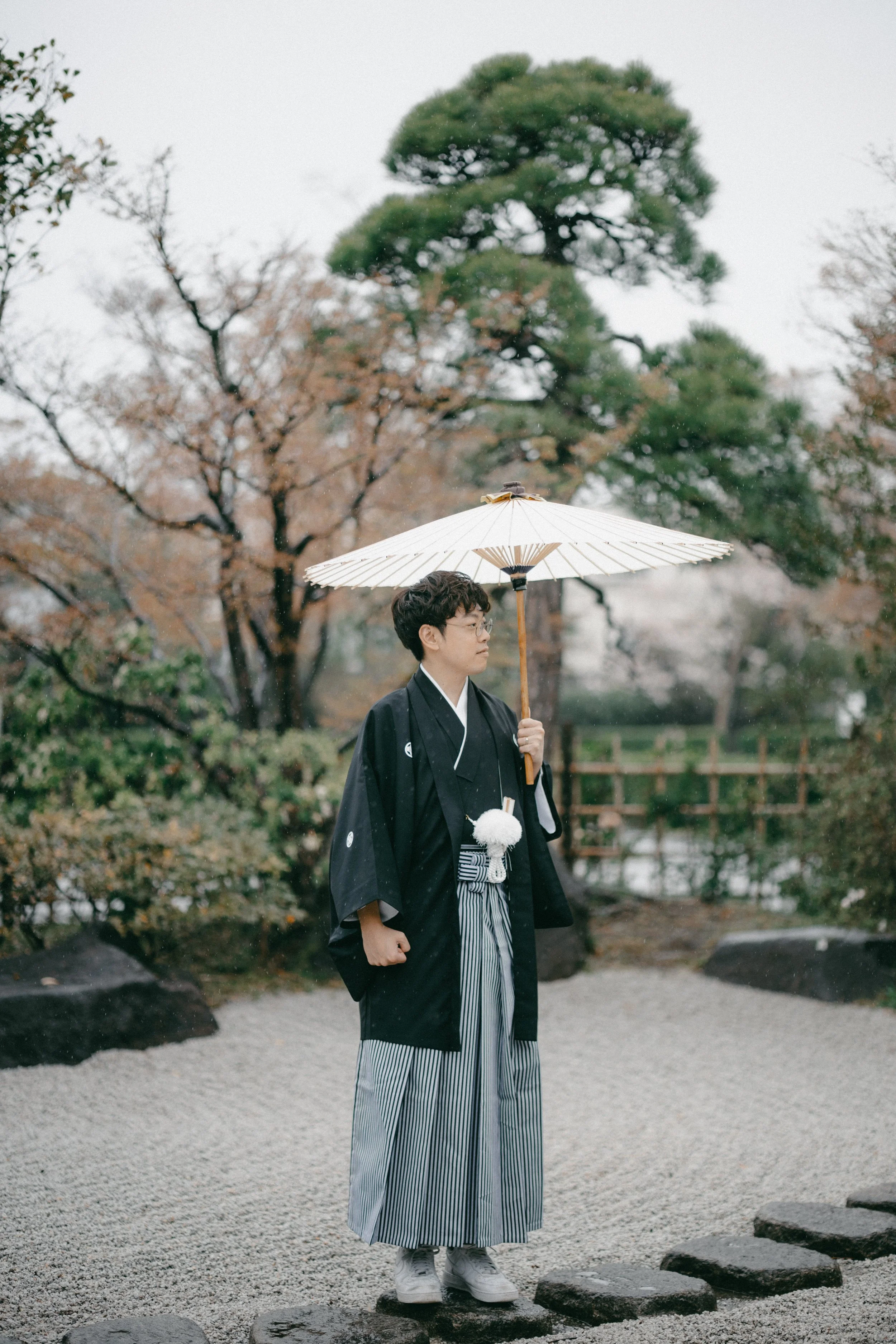 A young man dressed in traditional Japanese attire holding a paper umbrella outdoors in a garden with trees and rocks.