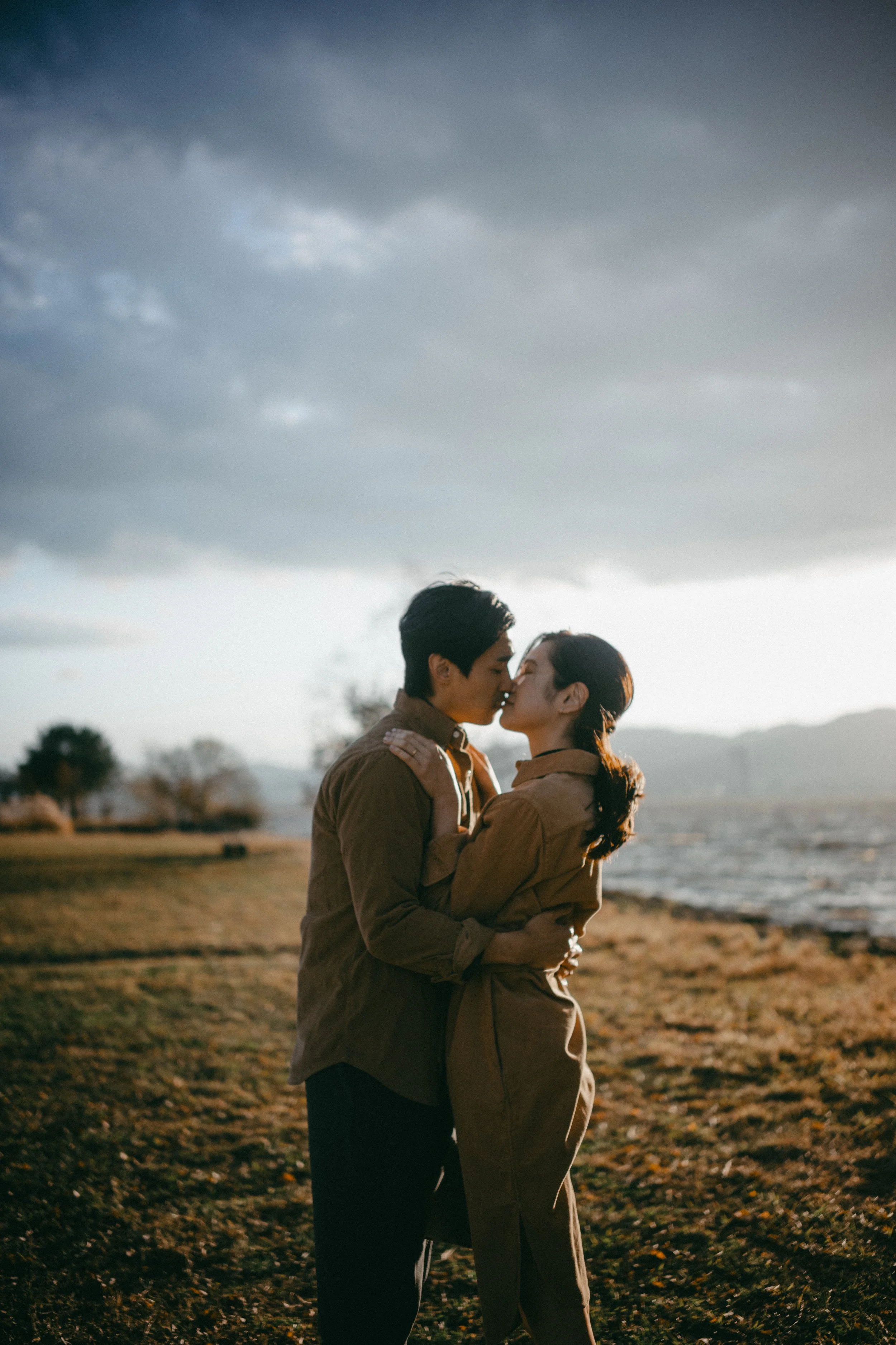 A couple sharing a kiss outdoors near a body of water during sunset.