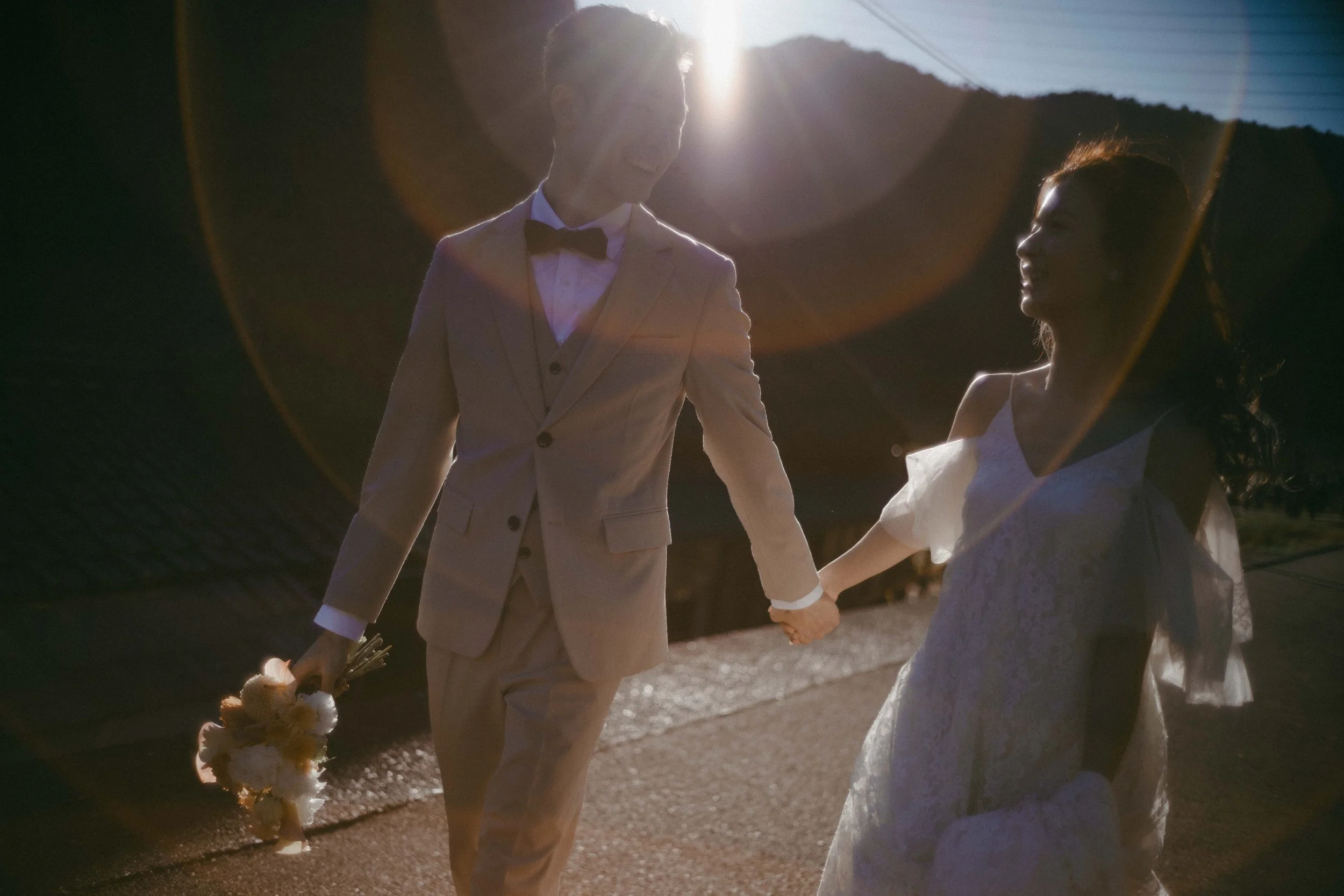 A newlywed couple holding hands outdoors during sunset, with the groom in a light-colored suit and the bride in a white dress, holding a bouquet of flowers.