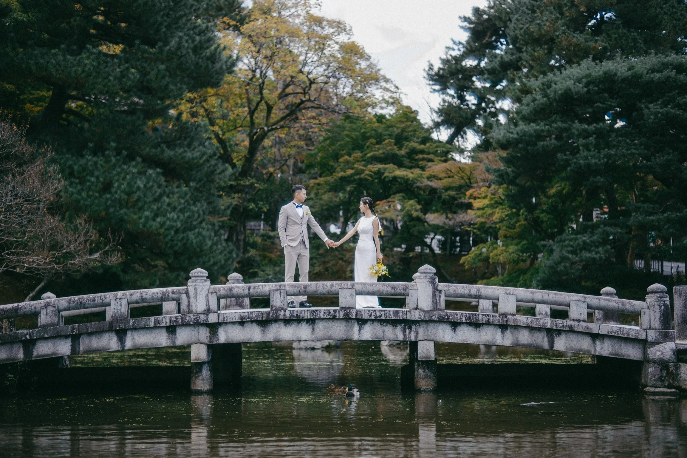 A bride and groom holding hands on a stone bridge over a pond in a park, surrounded by green trees.