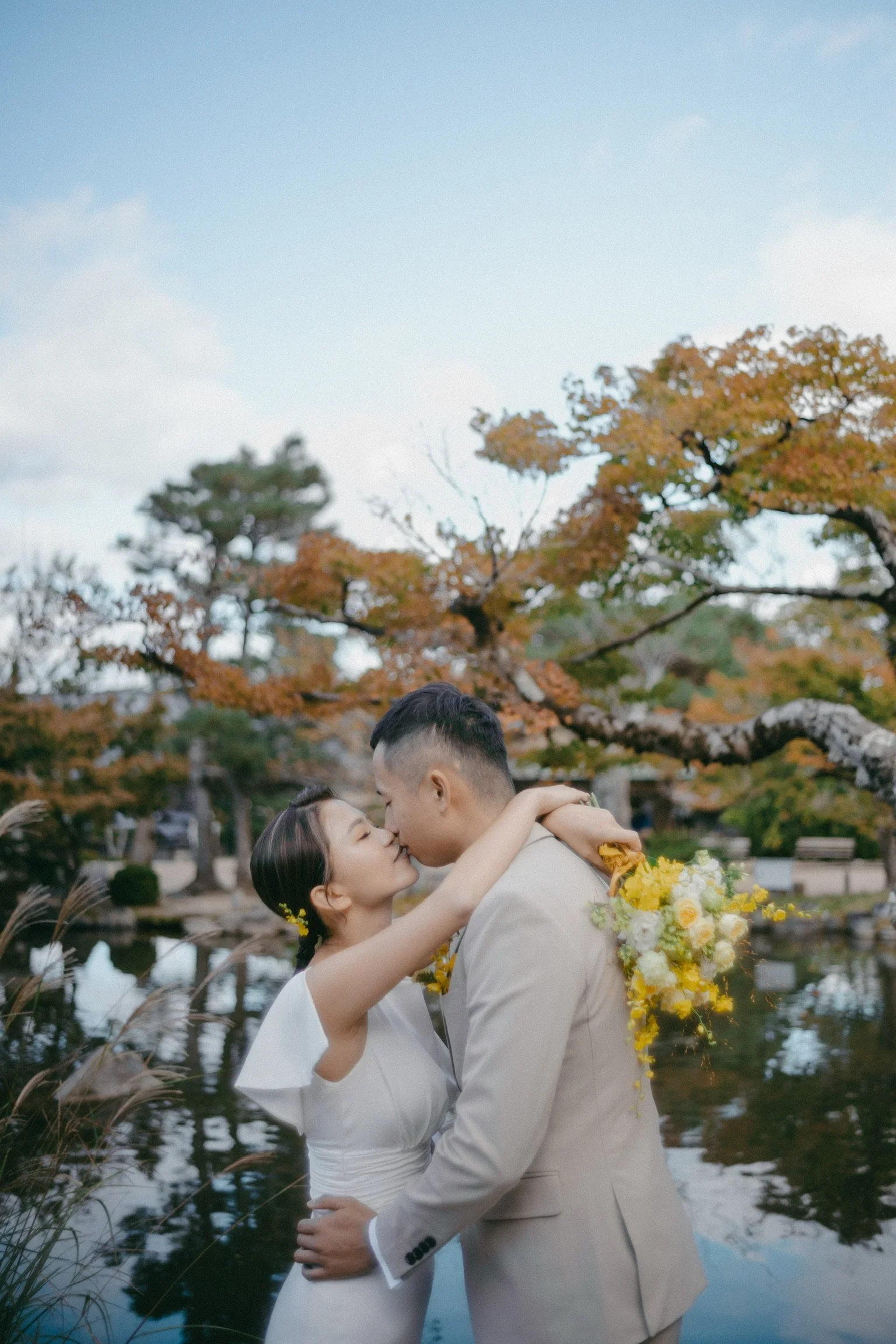 A couple sharing a kiss outdoors by a pond in a park with autumn foliage.