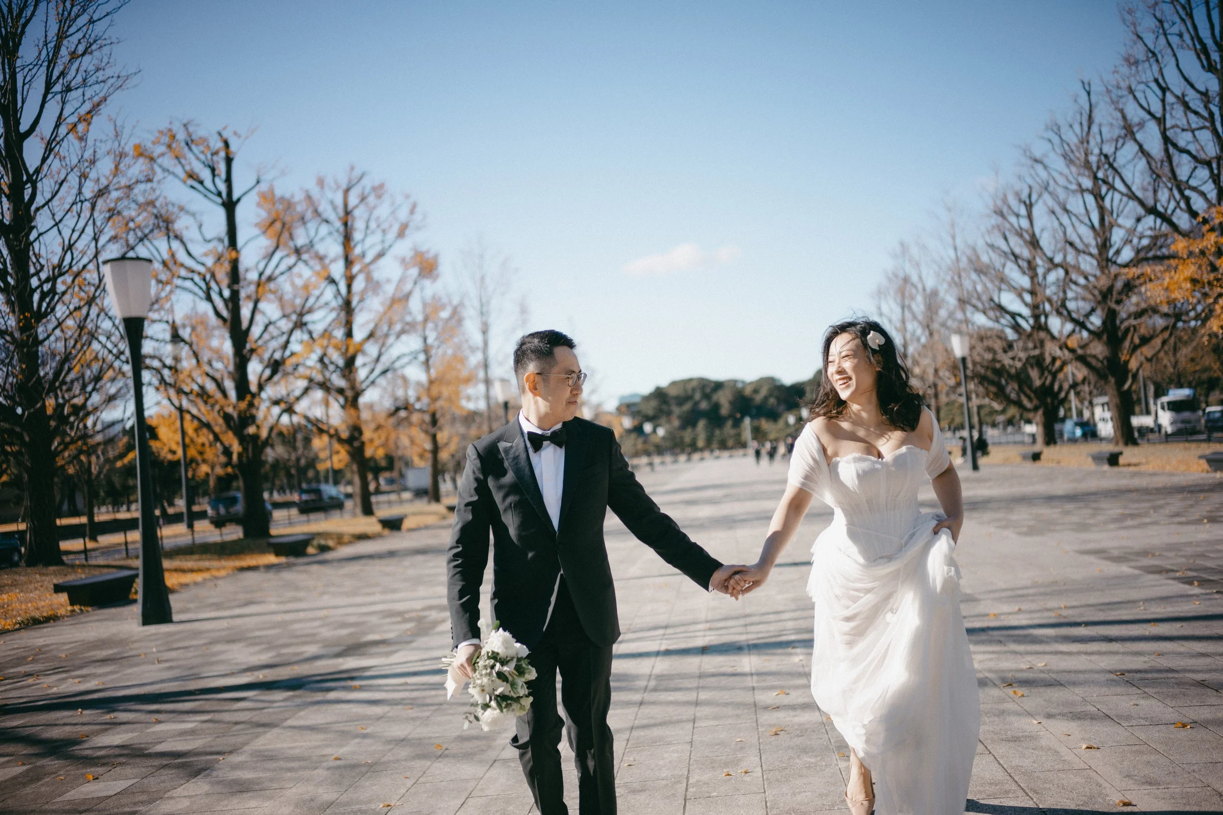 A bride and groom holding hands and walking in a park with leafless trees and clear blue sky, the groom in a black tuxedo and the bride in a white wedding dress carrying a bouquet of flowers.