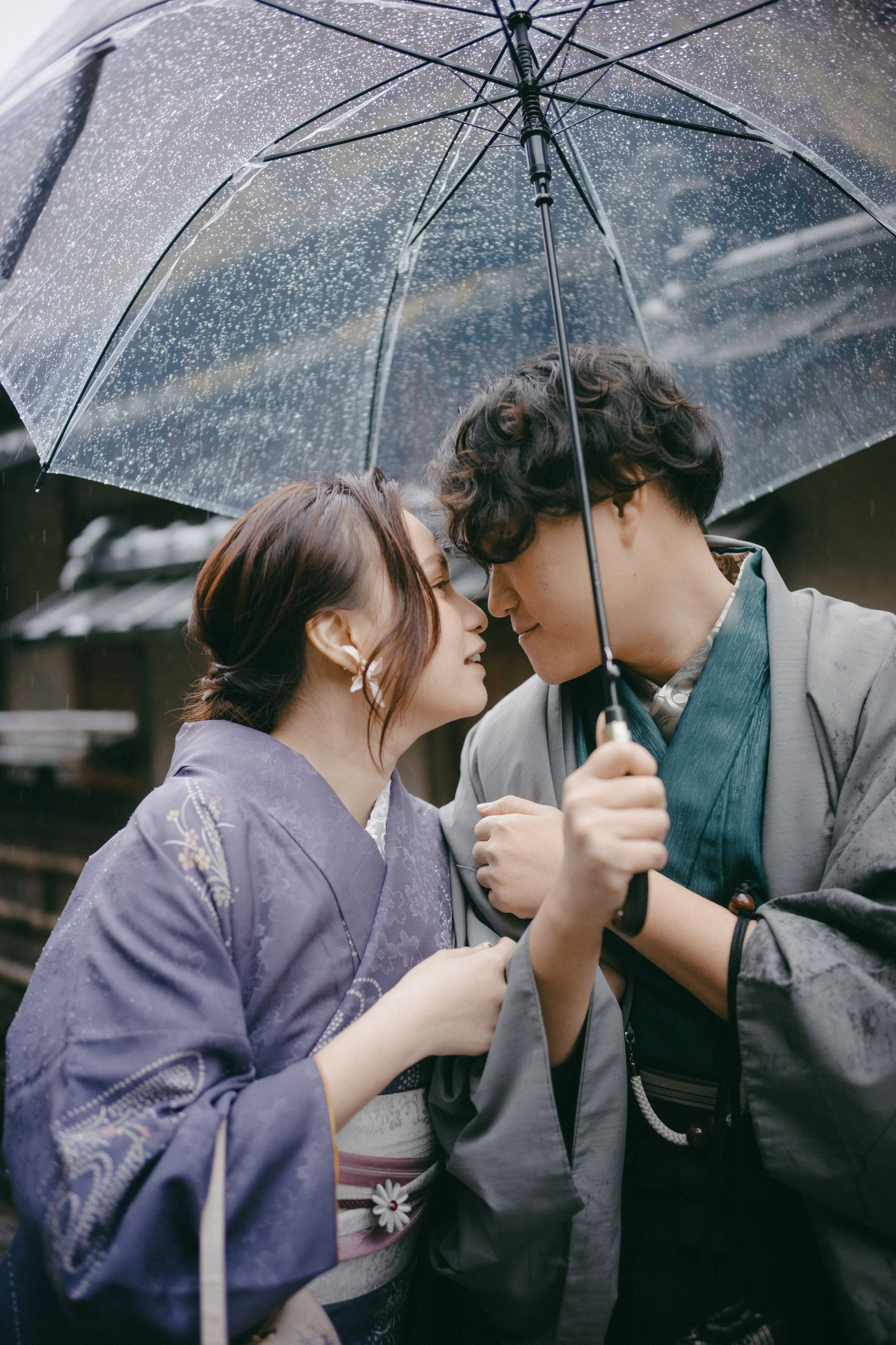 A couple dressed in traditional Japanese kimonos standing close under a transparent umbrella with raindrops on it, smiling and leaning their foreheads together.