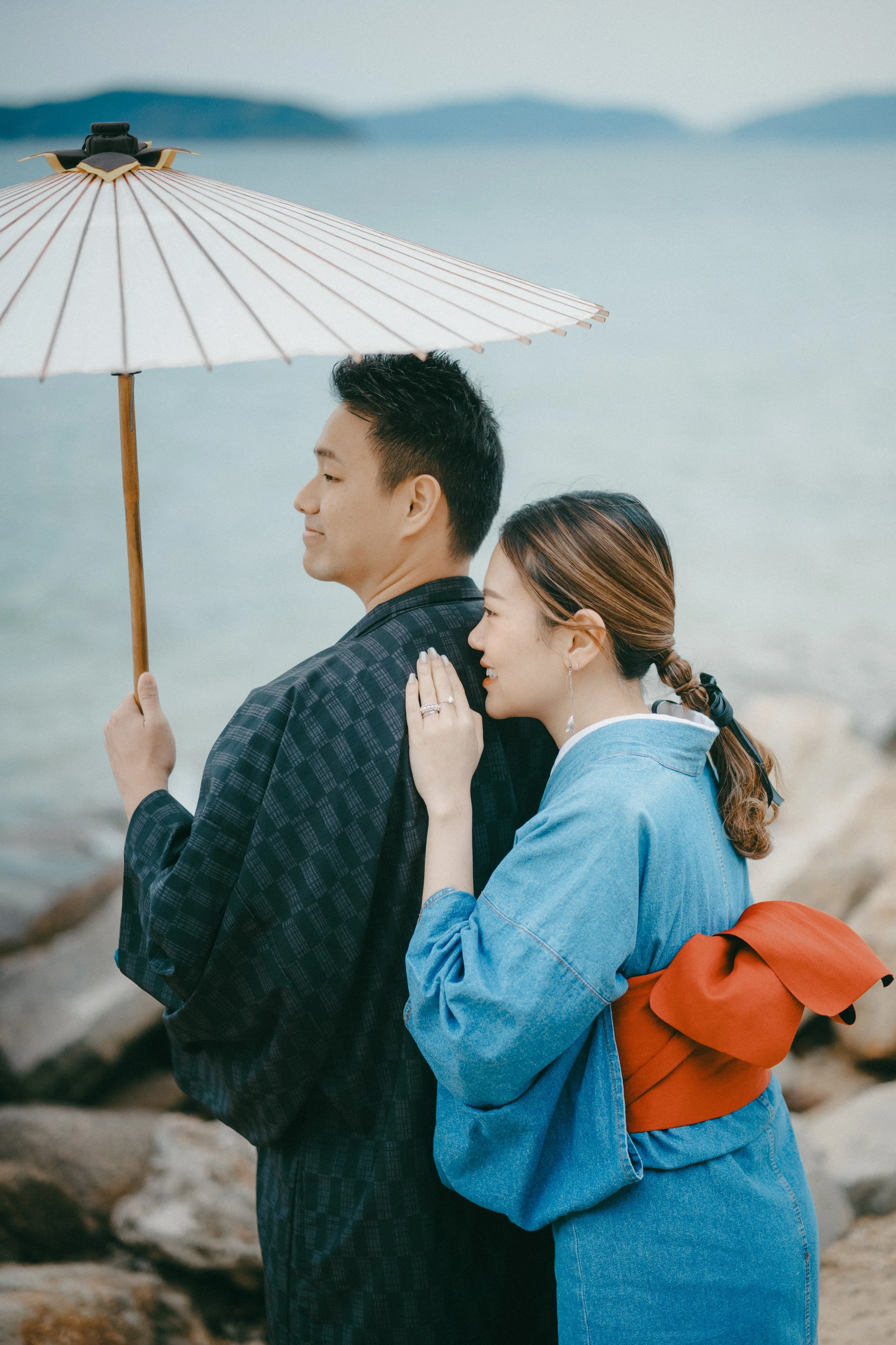 A couple at the beach, with the woman smiling and leaning on the man's shoulder, holding an umbrella over him. Rocks and water are visible in the background.