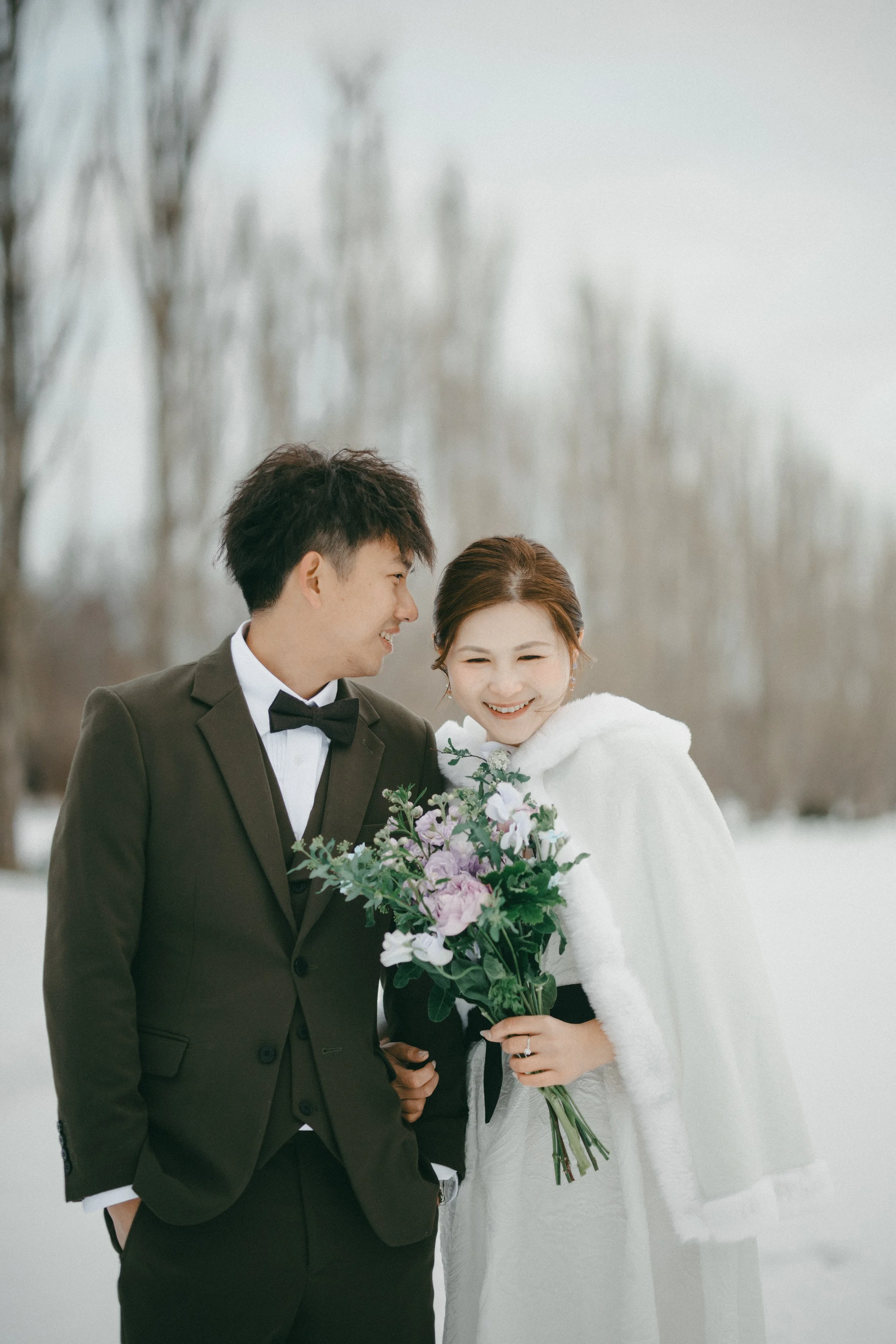 A happy couple in wedding attire standing outdoors in winter. The groom is wearing a black suit with a bow tie, and the bride is wearing a white dress with a white hooded cape. The bride is holding a bouquet of purple and white flowers, and they are 