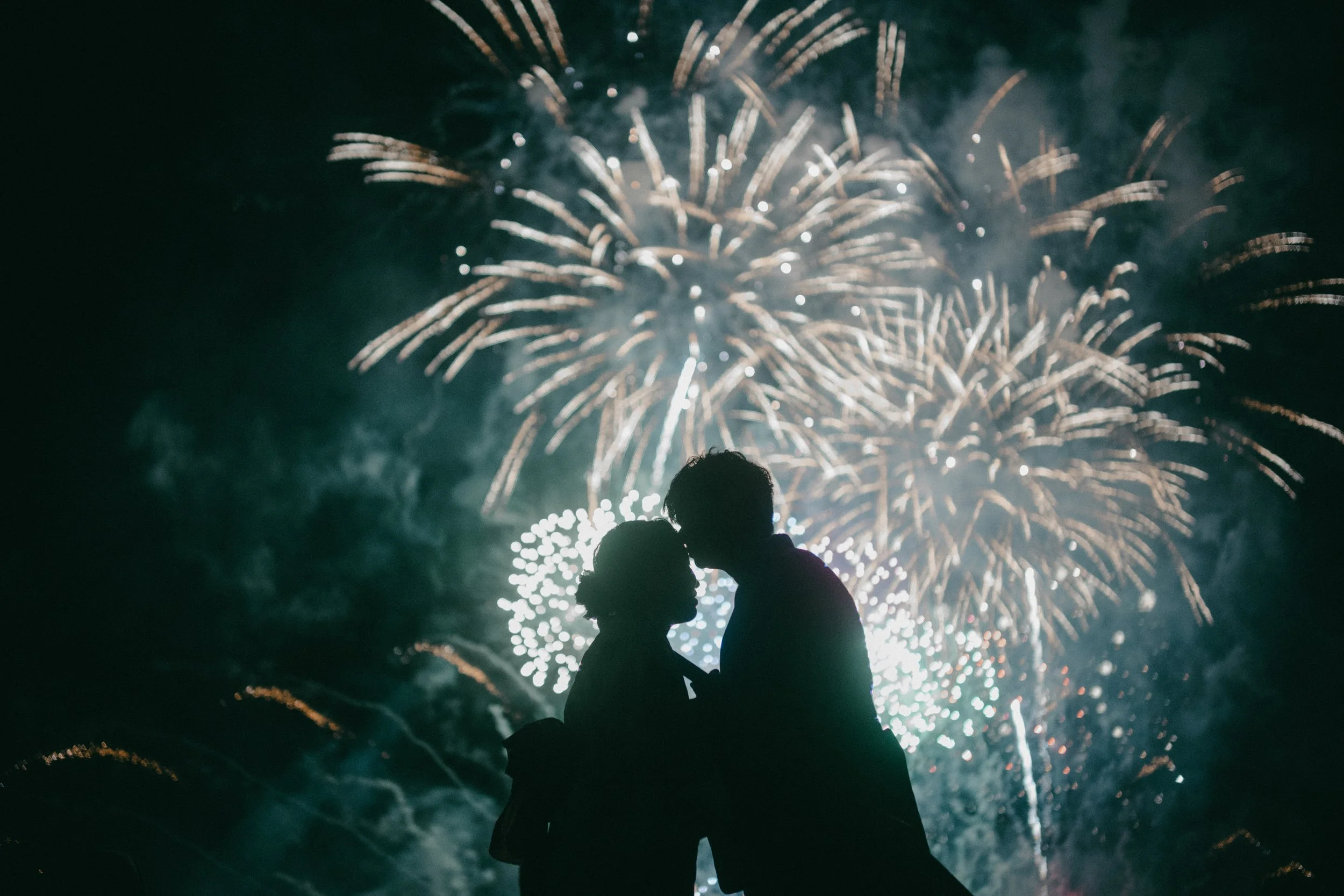 Silhouettes of a couple kissing or holding each other against a backdrop of colorful fireworks in the night sky.