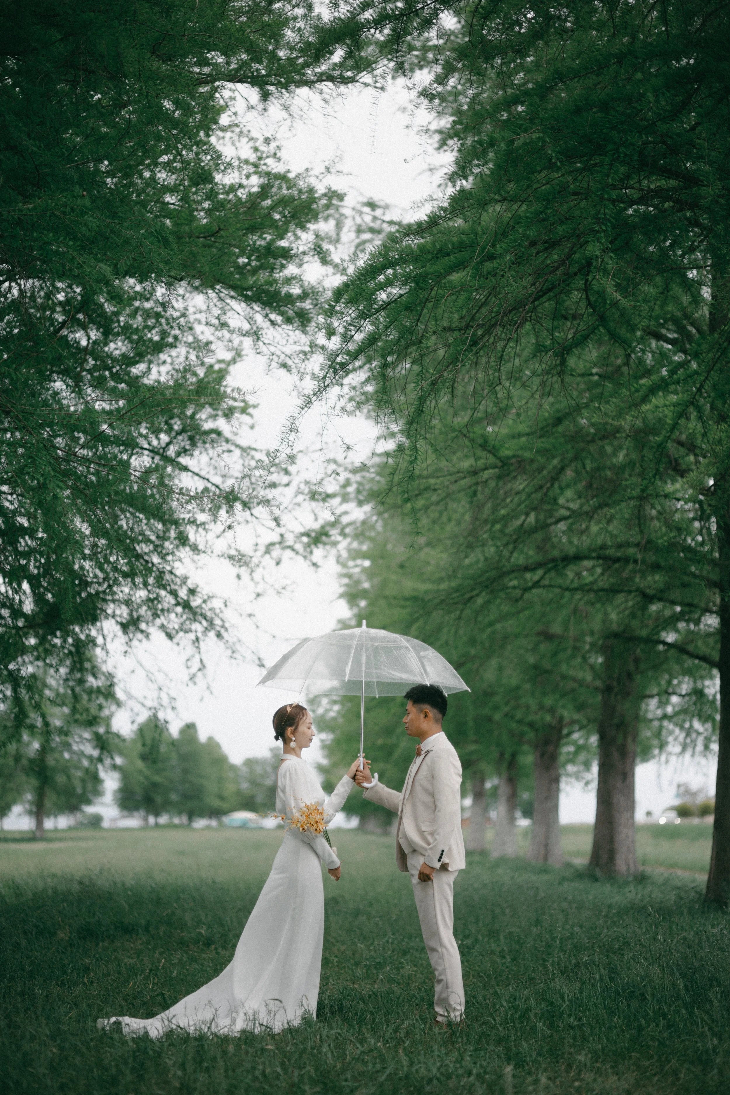 Bride and groom standing under a transparent umbrella in a park, surrounded by green trees, during overcast weather.