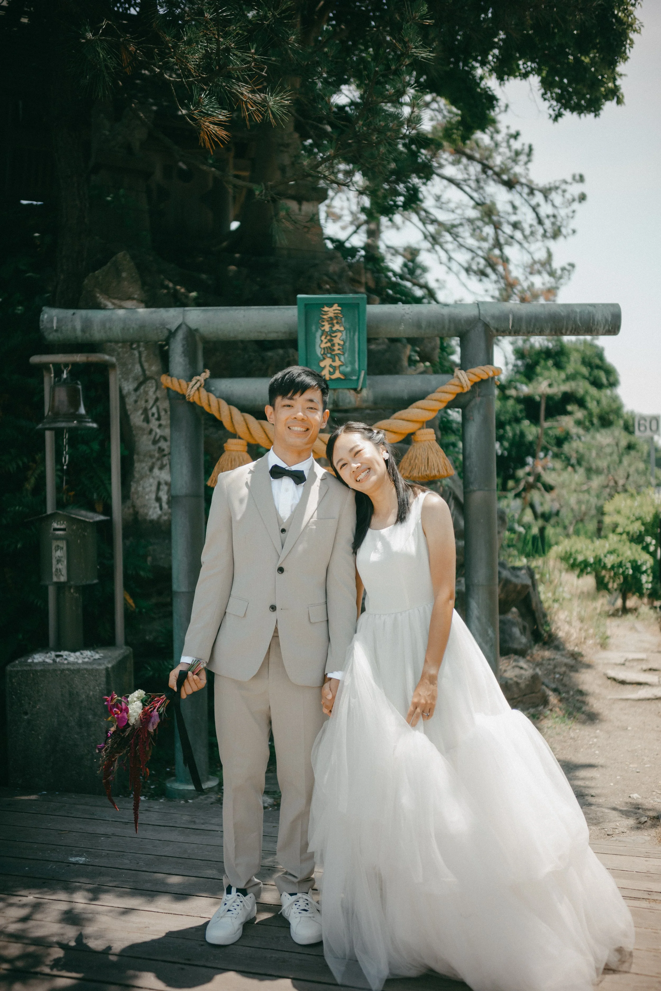 A happy bride and groom in wedding attire standing outdoors in front of a traditional Japanese torii gate, smiling and holding hands, with trees and greenery surrounding them.