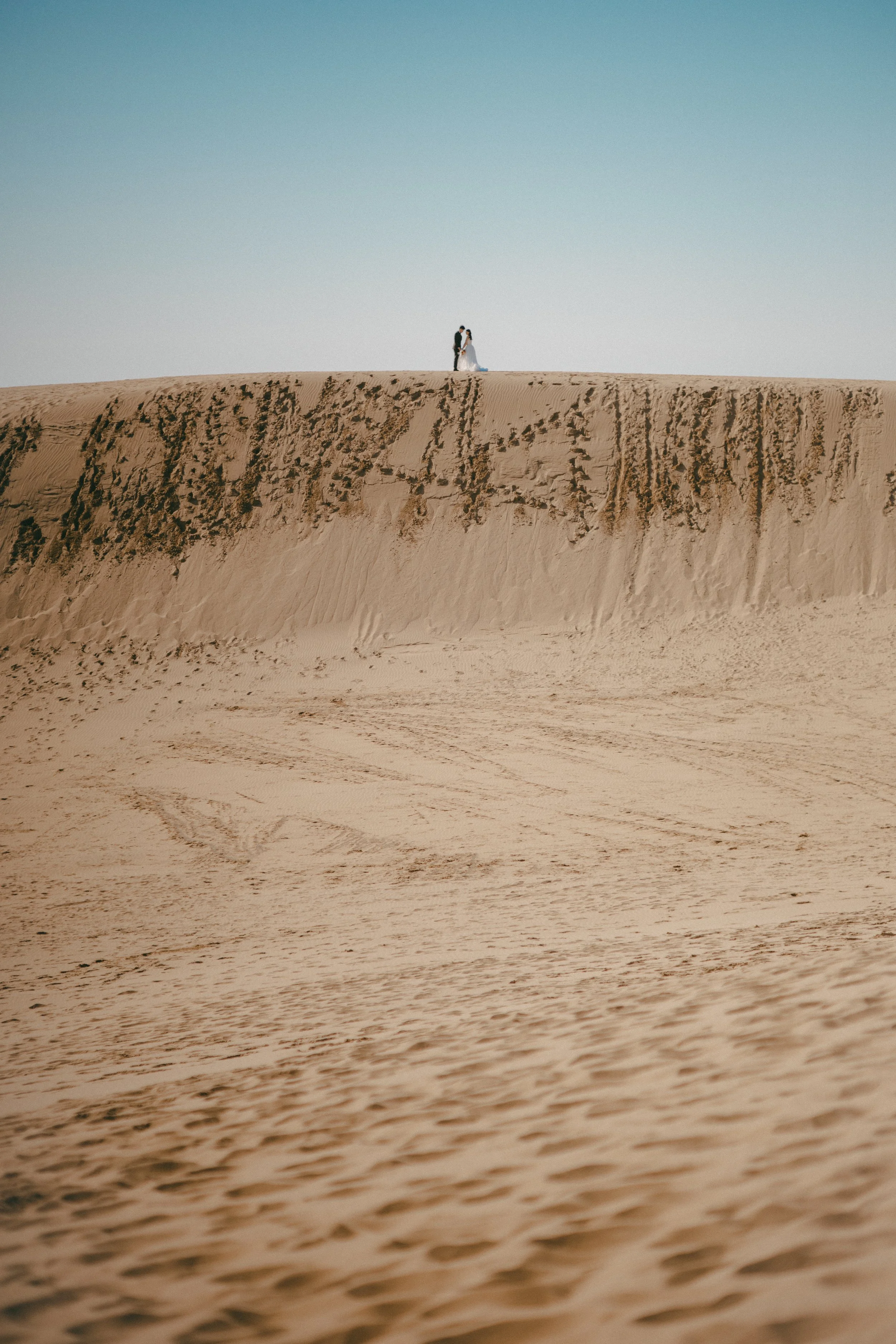 A bride and groom stand on top of a sand dune, with the desert terrain in the foreground and clear blue sky above.