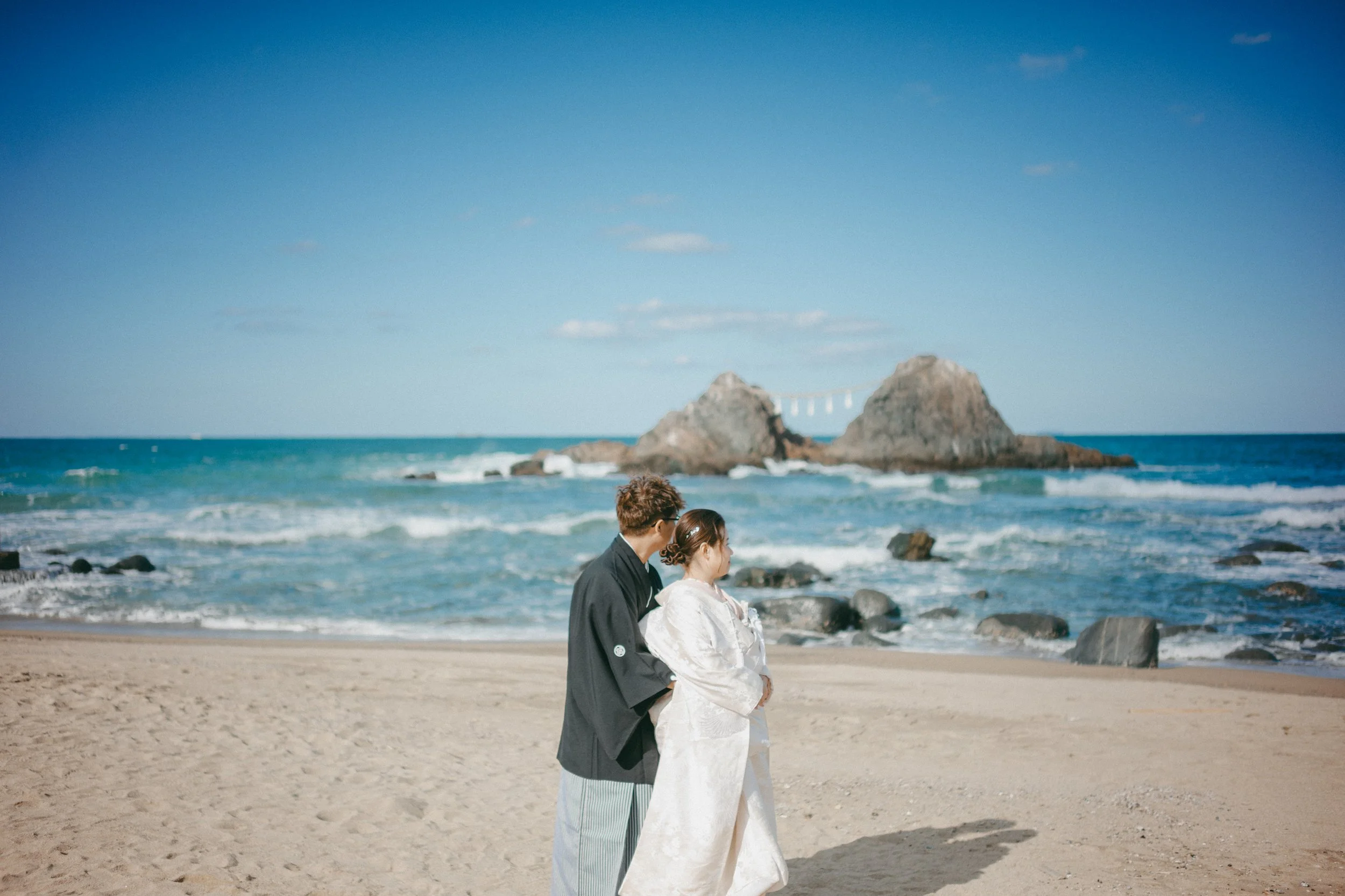 A couple dressed in traditional Japanese wedding attire standing on a sandy beach with the ocean and two large rocks in the background.
