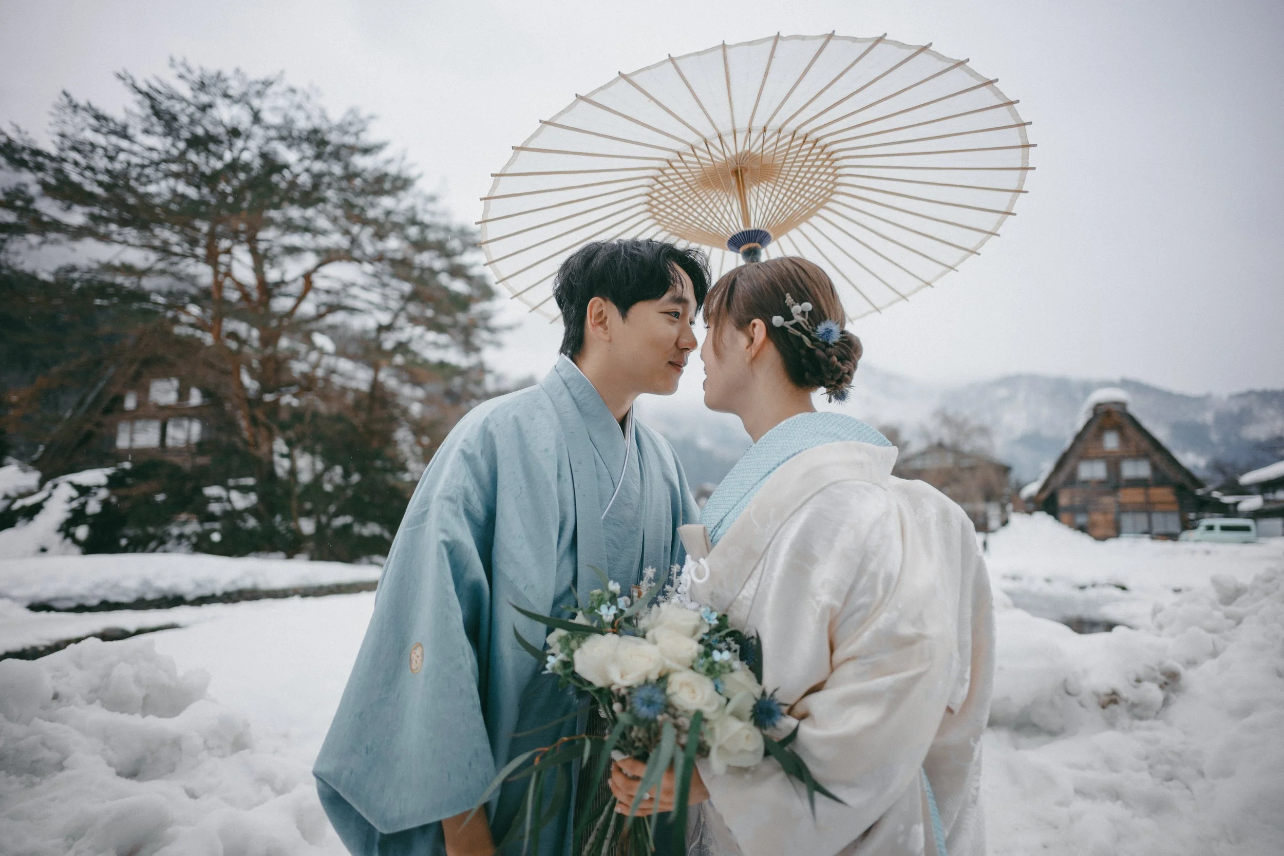 A couple dressed in traditional Japanese wedding attire stands close together in a snowy landscape, holding a bouquet of white roses and blue flowers, with a large umbrella overhead, and traditional houses in the background.