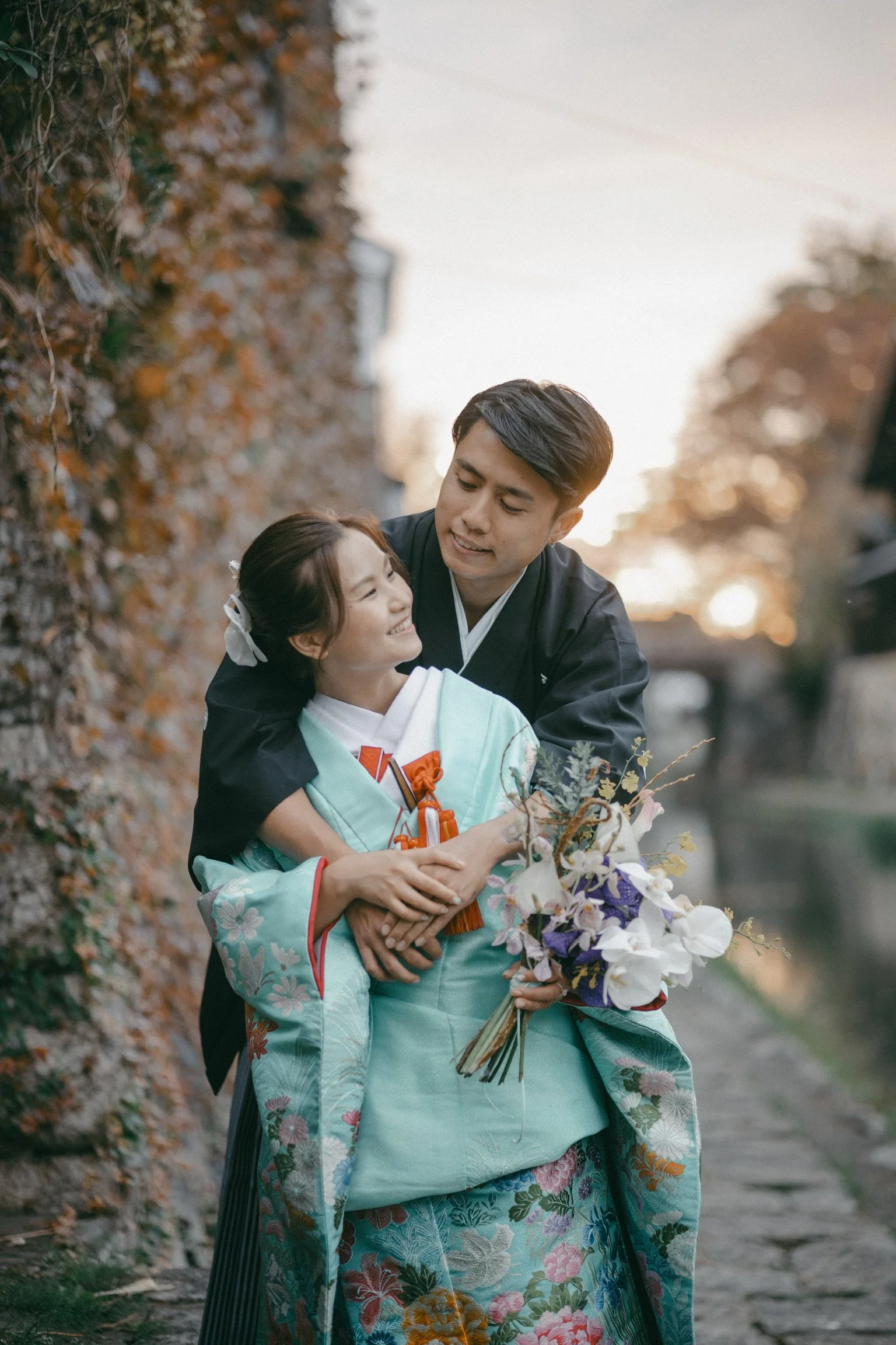 A Japanese couple in traditional kimono celebrating outdoors at sunset, with the woman holding a bouquet of flowers and smiling as the man embraces her.