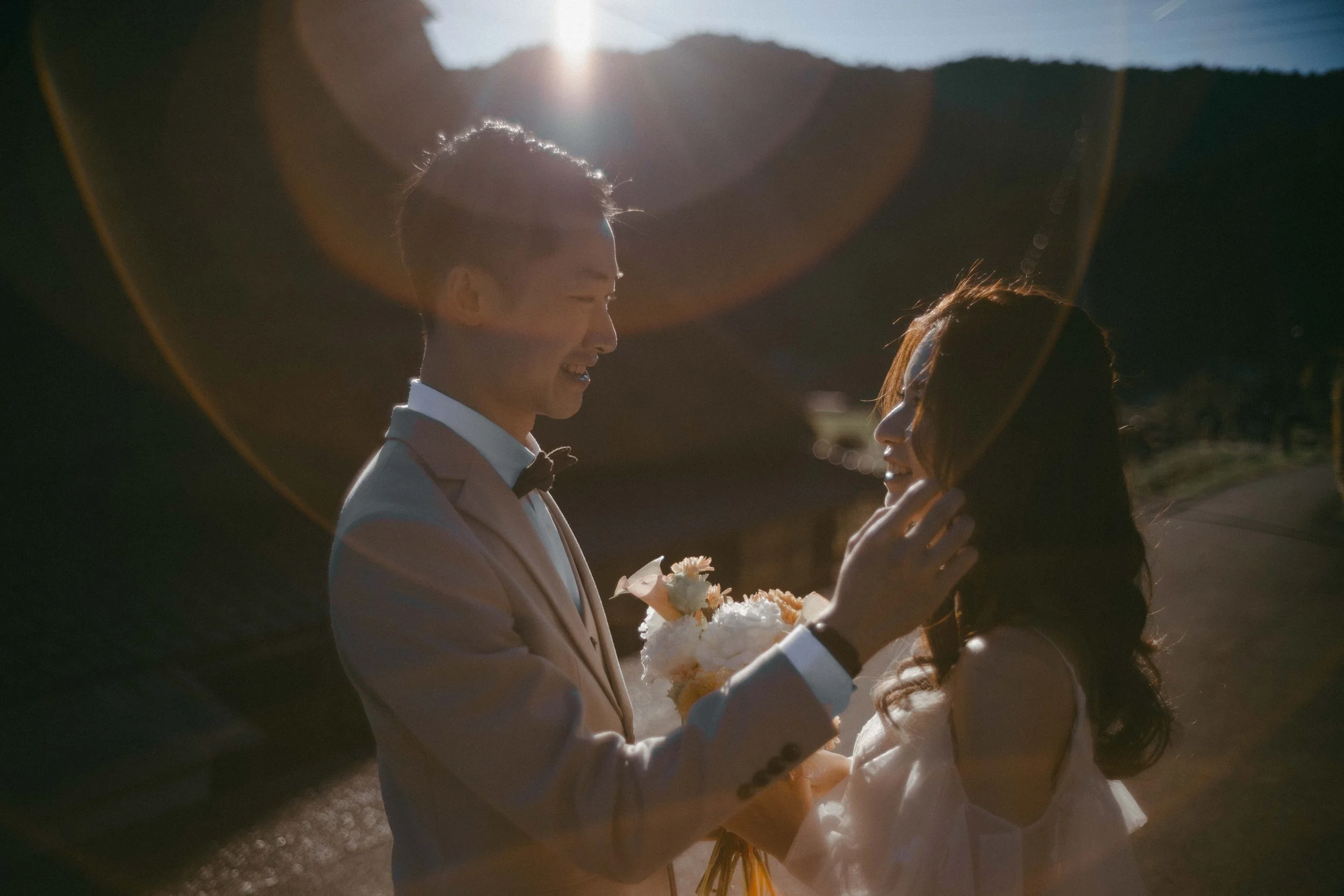A groom and bride sharing a moment outdoors during sunset, with the groom holding a bouquet of flowers and touching the bride's face, both smiling.