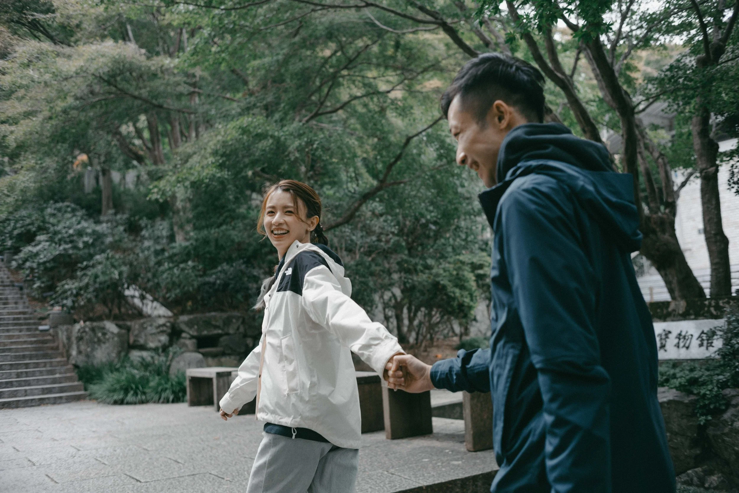 A woman and a man holding hands, smiling, outdoors among trees in a park.