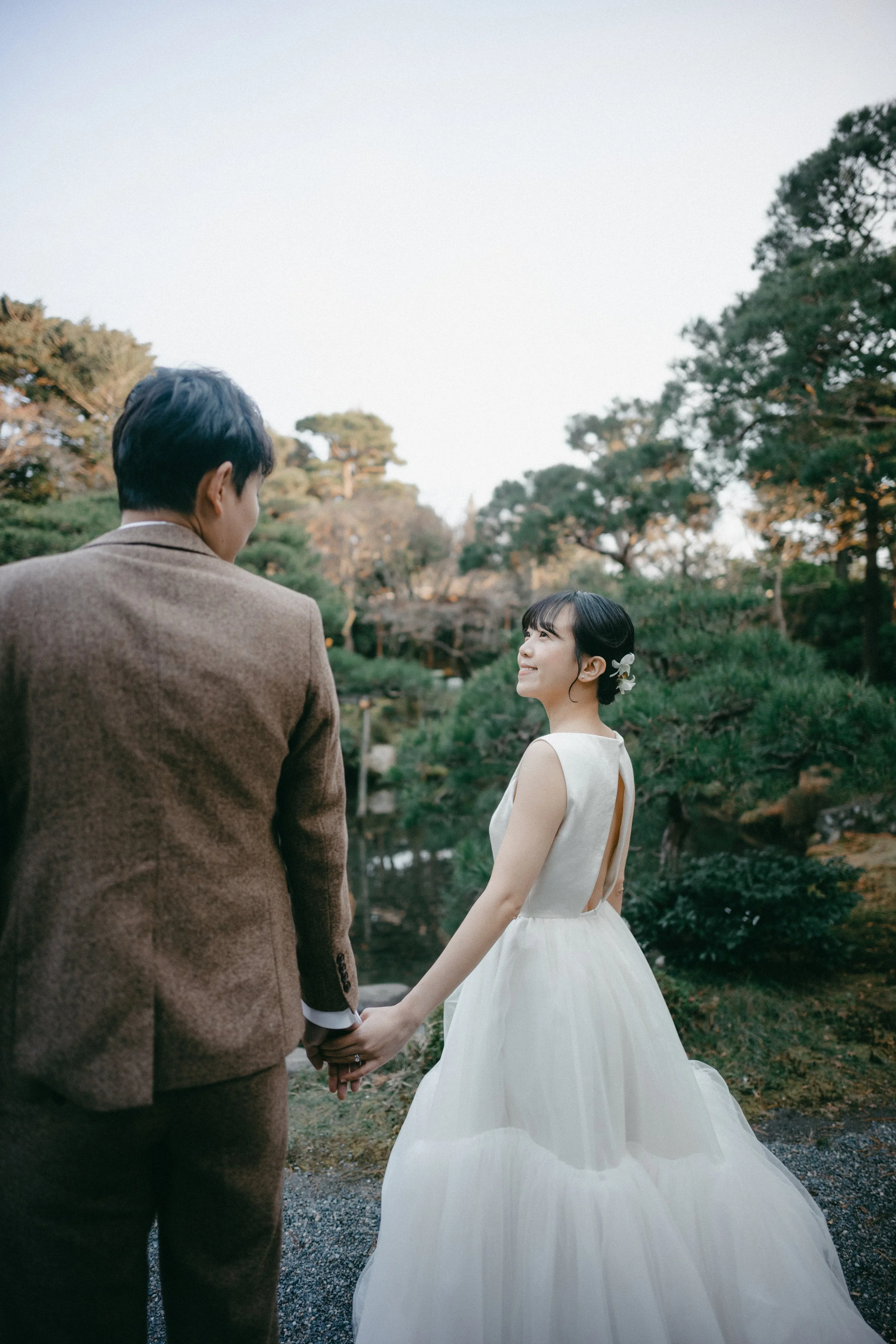 A bride and groom holding hands in a garden during sunset, with trees and a pond in the background.