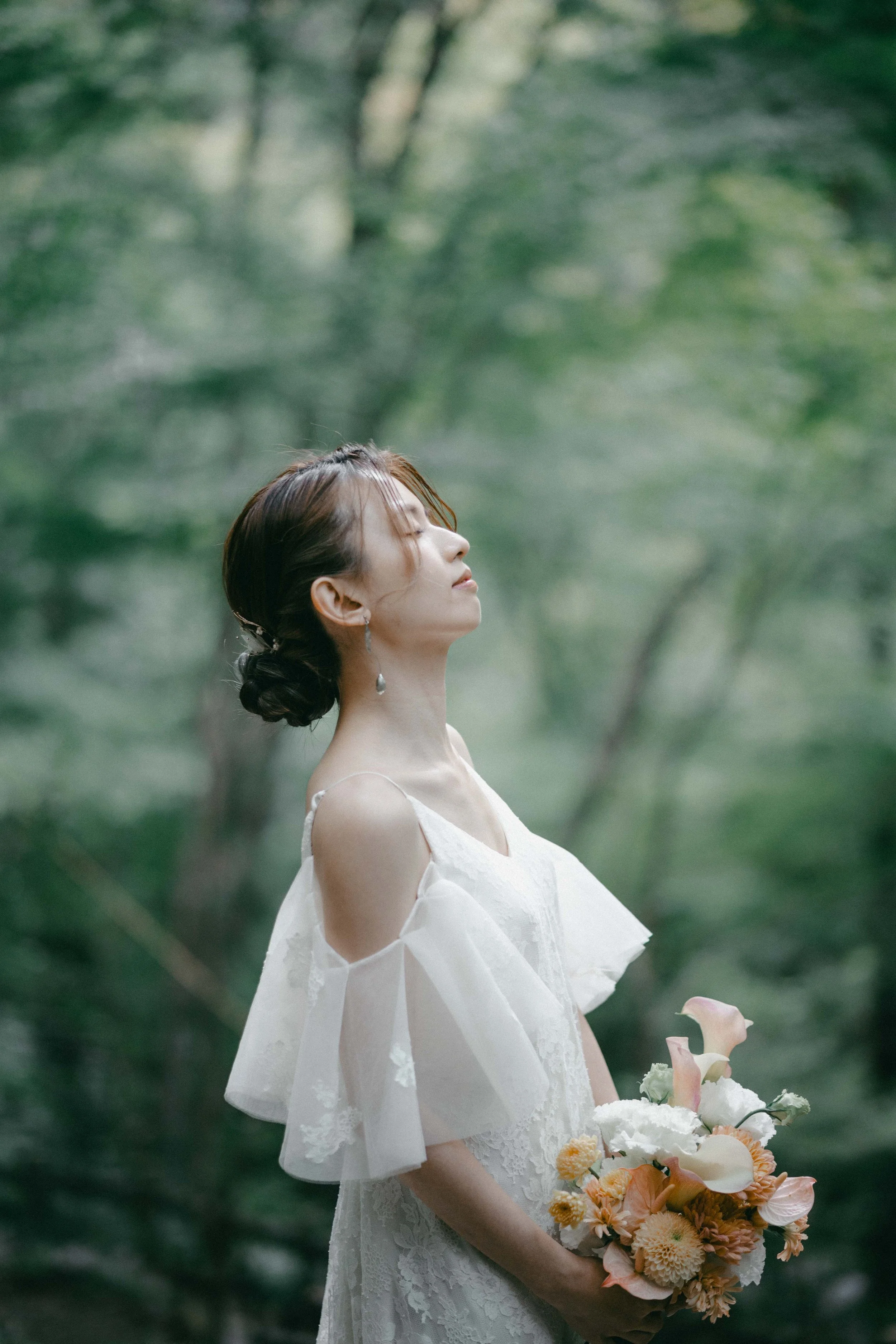 A woman in a white dress holding a bouquet of flowers standing in a green forest with her eyes closed and head tilted back.