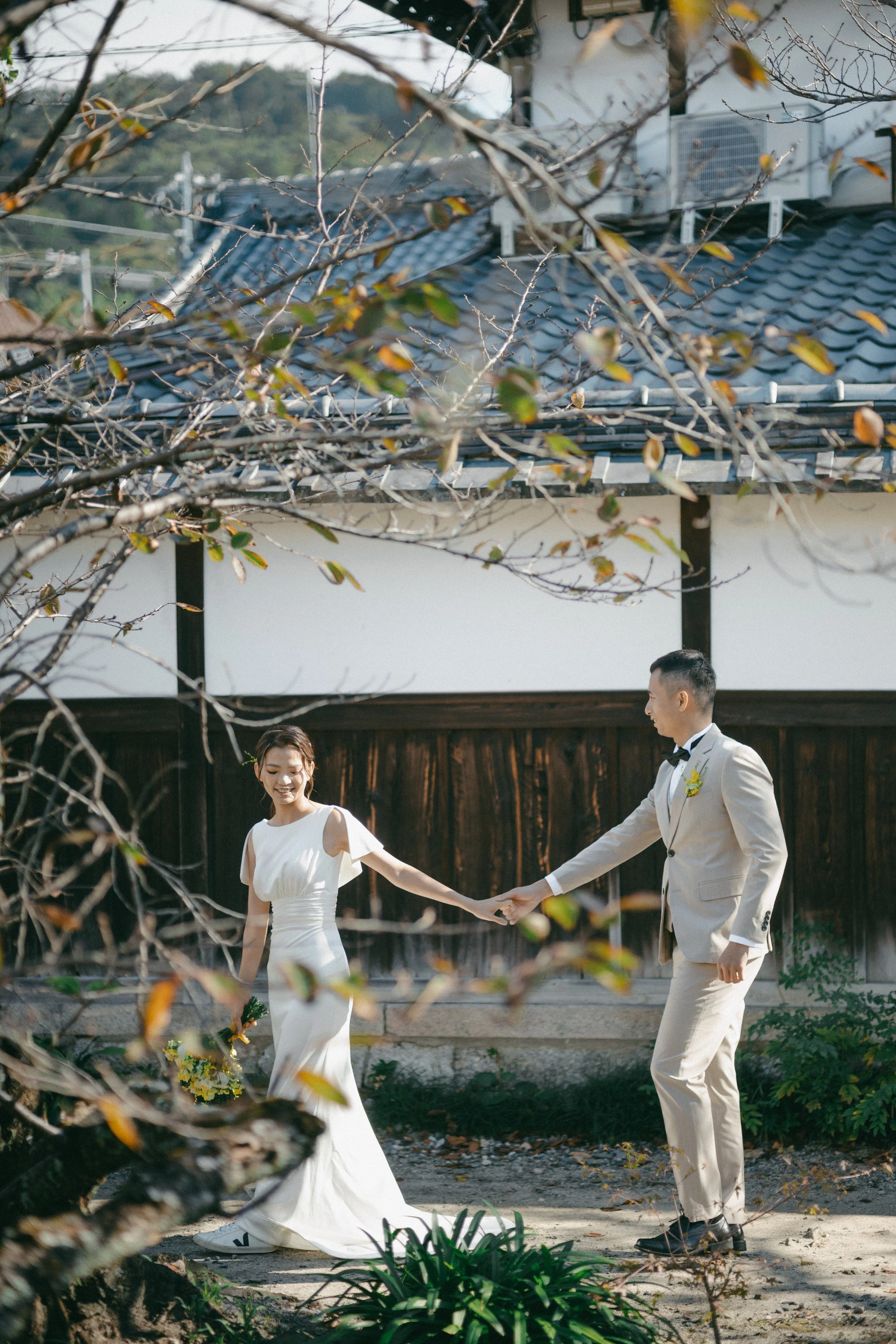 A bride and groom holding hands outdoors. The bride is wearing a white wedding dress and holding a bouquet. The groom is dressed in a cream suit with a black bow tie. They are walking on a path with trees and traditional Japanese-style buildings in t