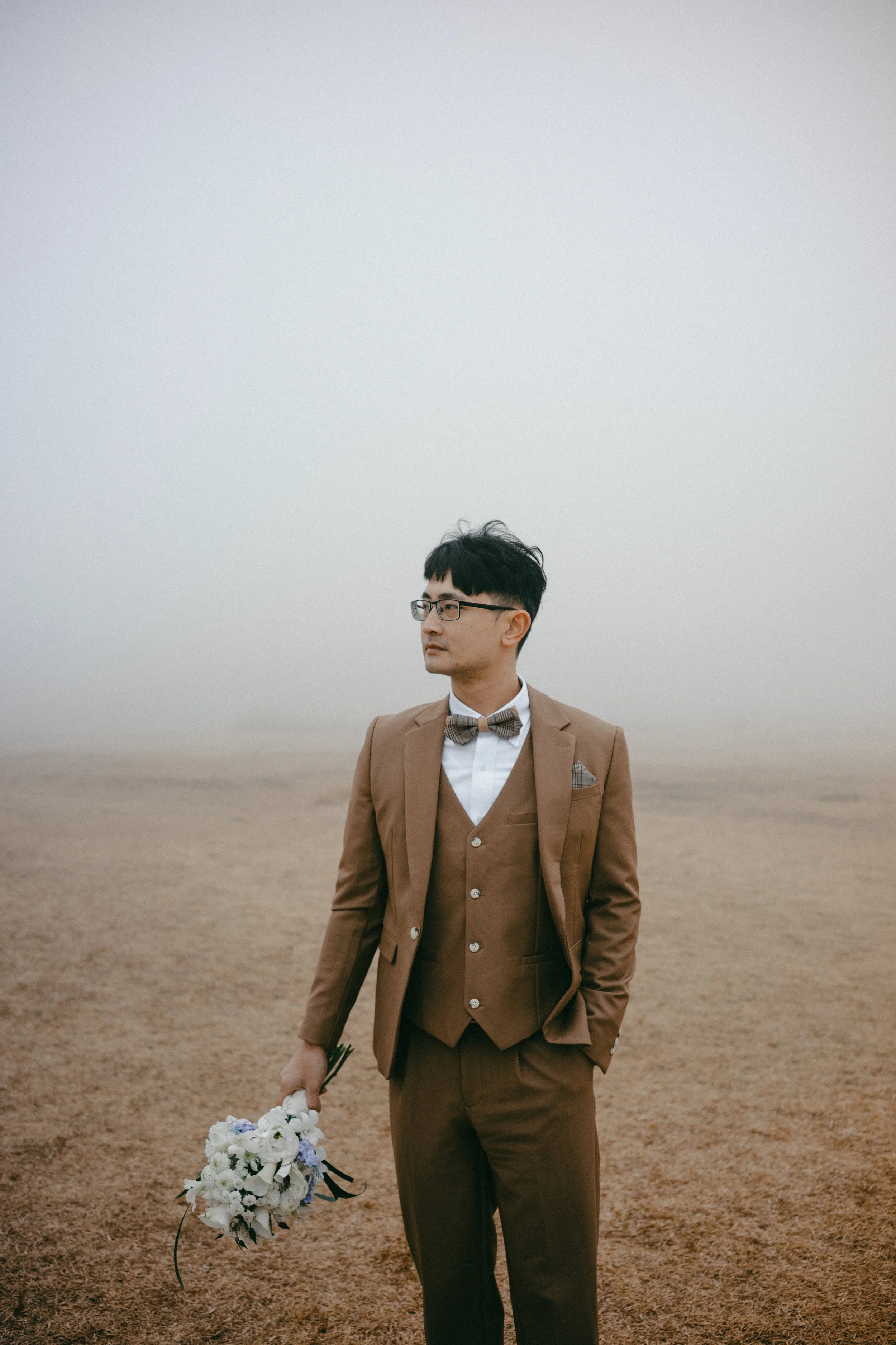 A man in a brown suit with a bow tie, holding a bouquet of flowers, standing on a foggy, barren landscape.