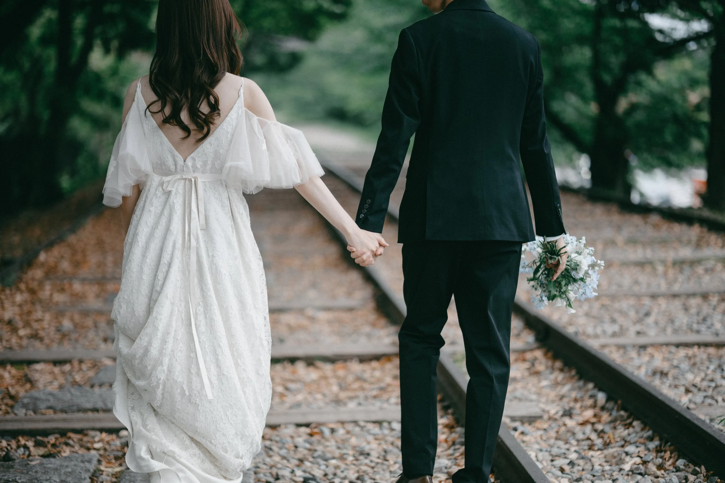 A bride and groom walking hand in hand along railroad tracks, with the bride wearing a white lace wedding dress with open shoulders and the groom in a black suit, holding a bouquet of flowers.