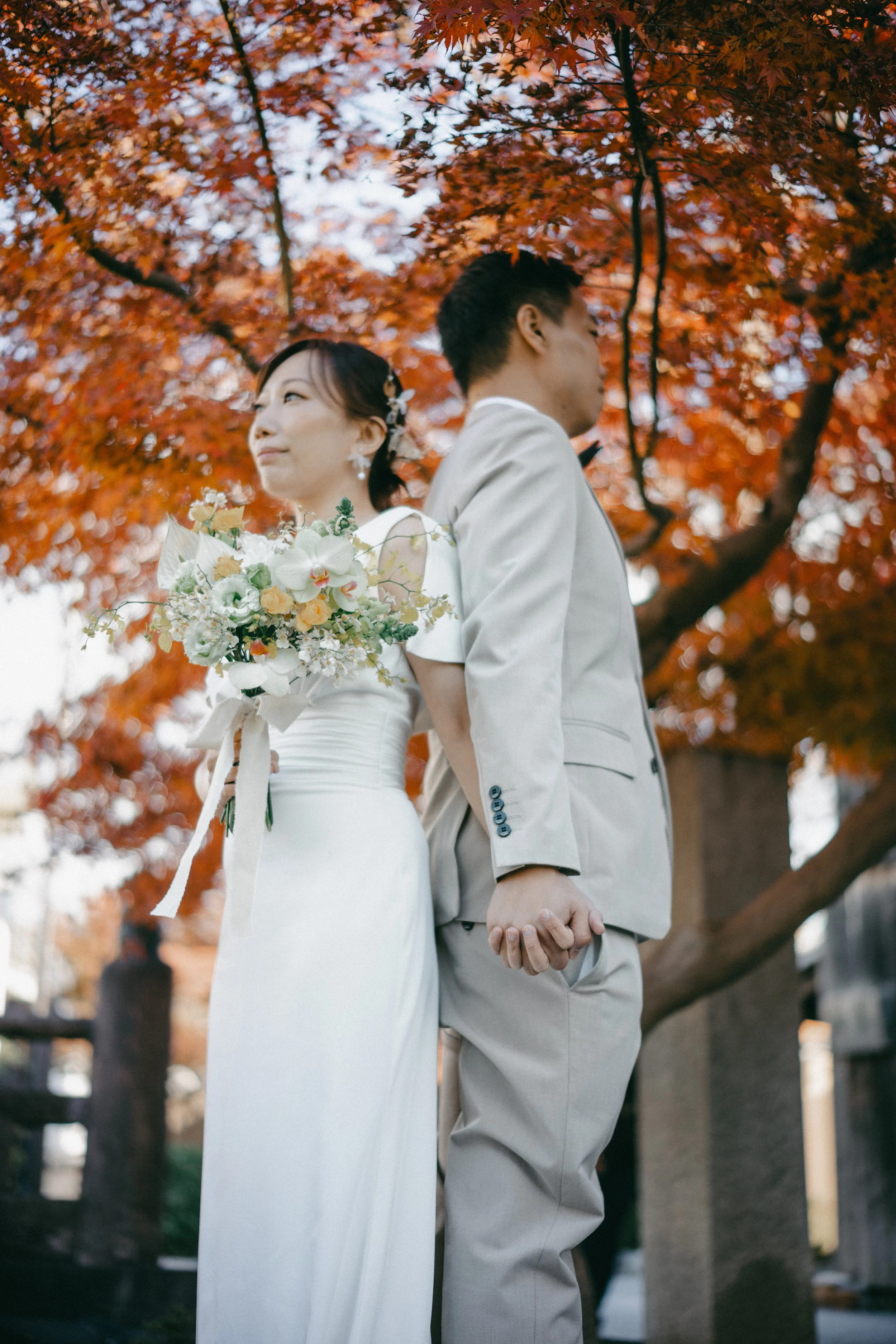 A bride and groom holding hands back-to-back outdoors during autumn, with colorful fall foliage in the background. The bride is holding a bouquet of flowers and wearing a white dress, while the groom is dressed in a light-colored suit.
