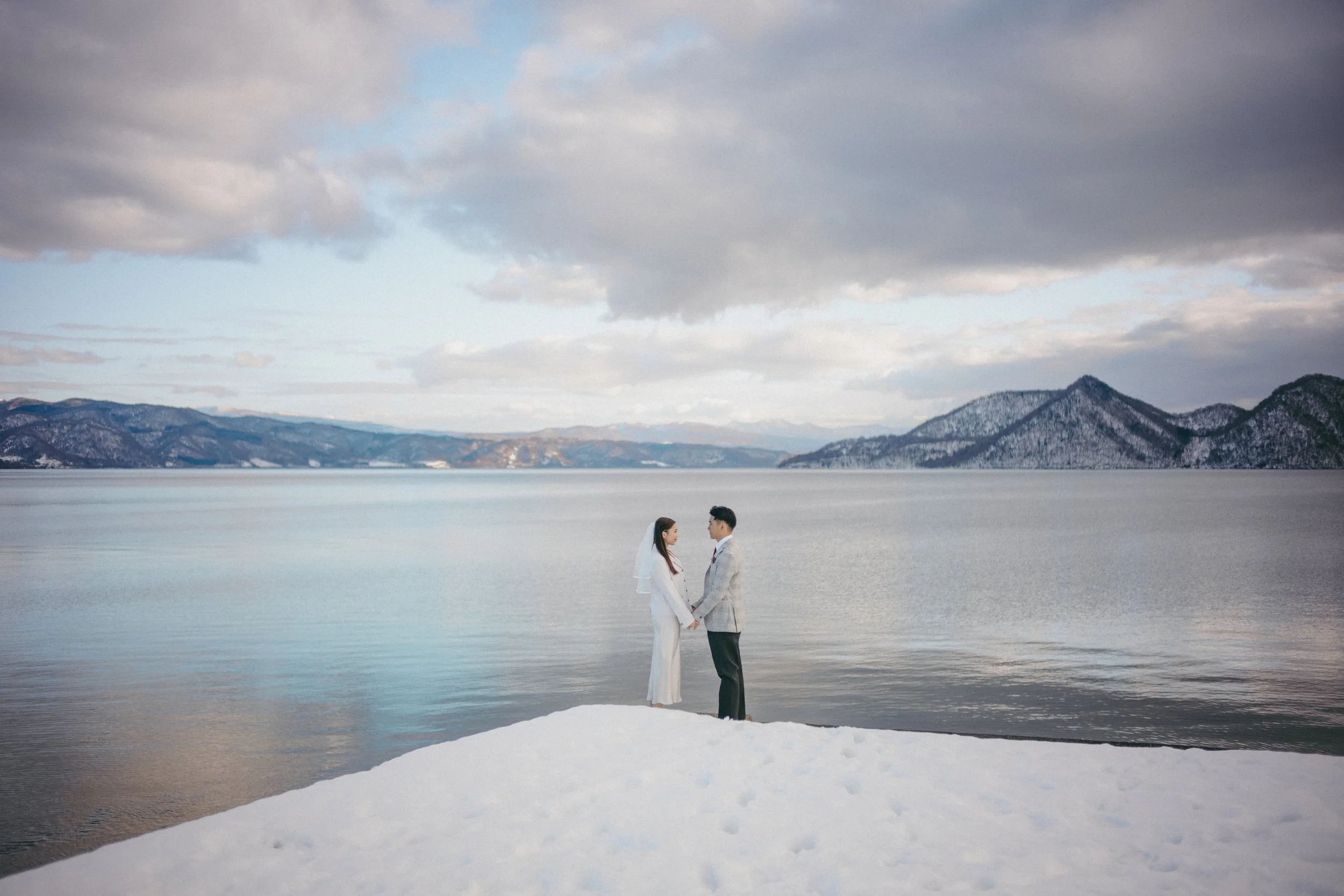 A bride and groom standing hand in hand on snow by a lake with mountains in the background.