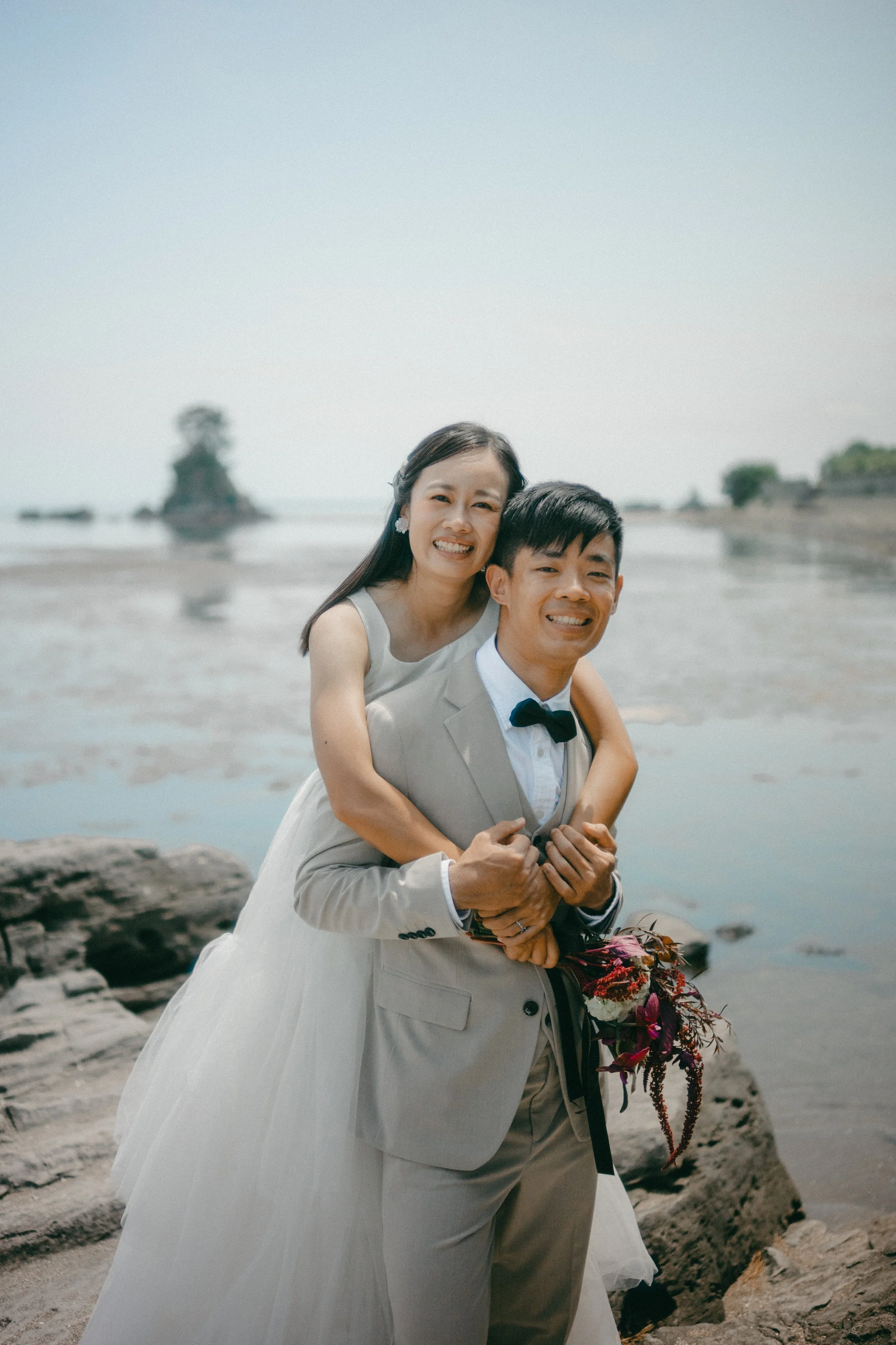 A smiling couple dressed in wedding attire, the woman in a white gown and the man in a light grey suit with a bow tie, standing on rocks by the water with a scenic background.