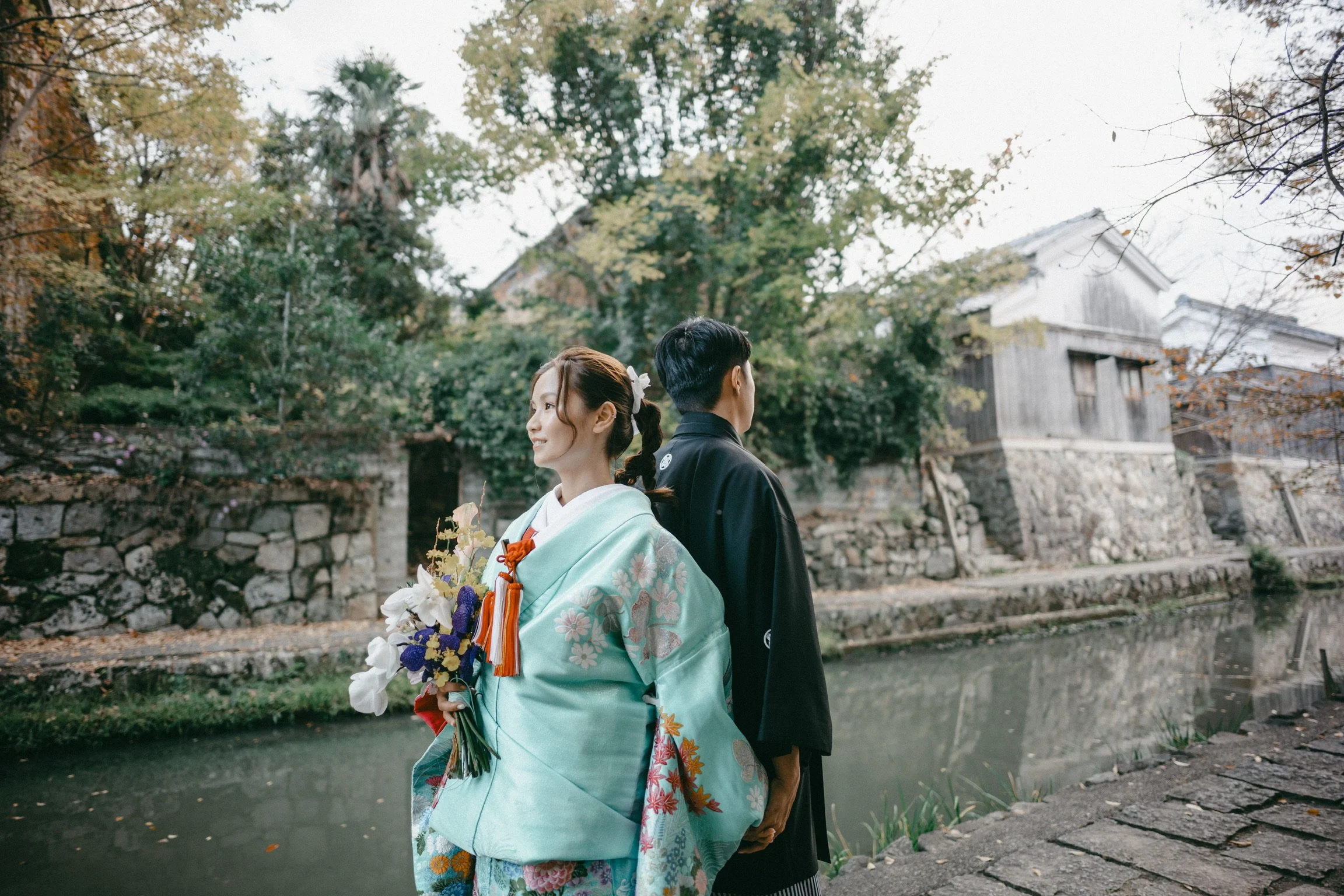 A woman and a man in traditional Japanese attire standing by a river, with trees and old buildings in the background.