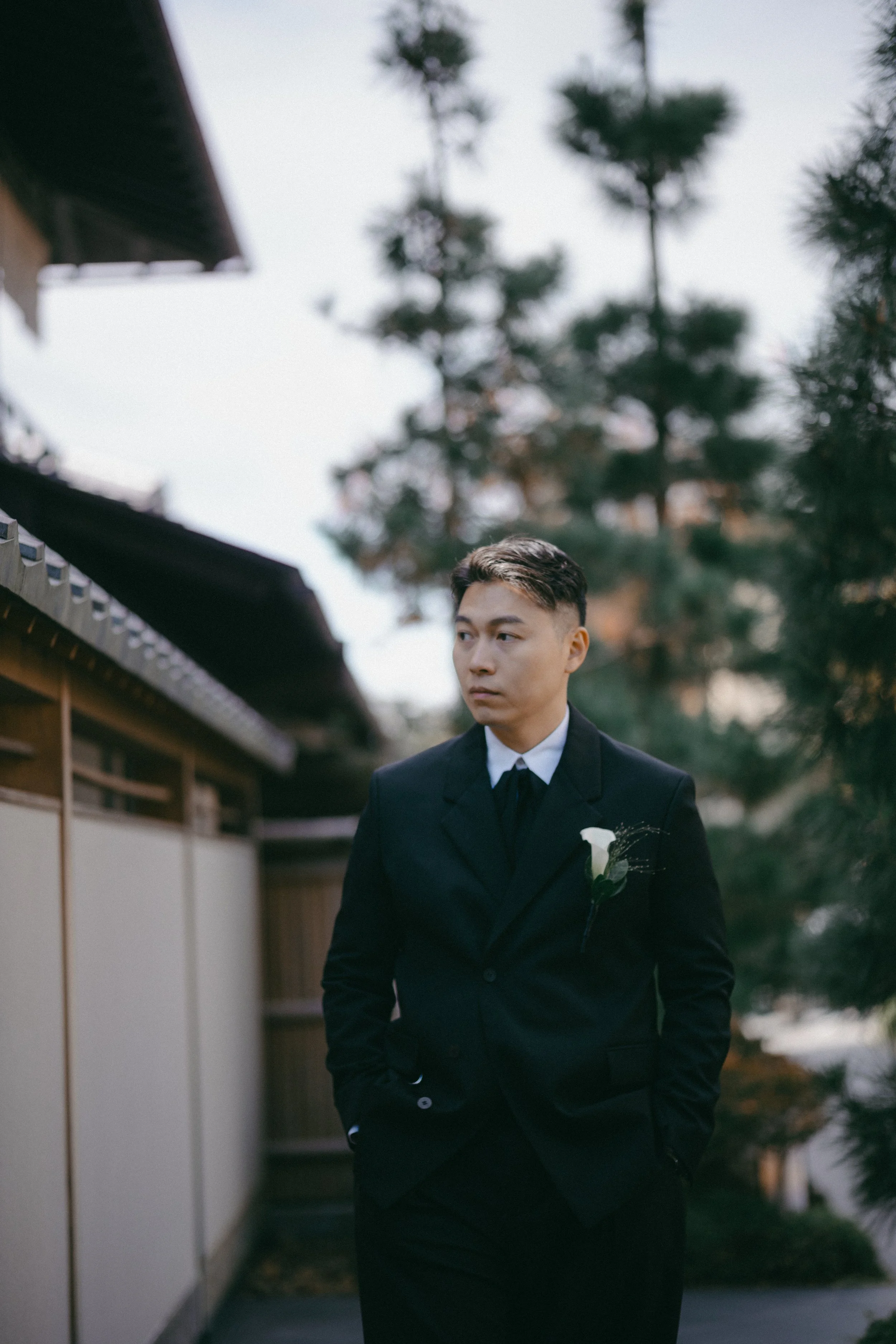 A young man in a black suit with a white boutonniere walking outdoors near a wooden fence and trees.