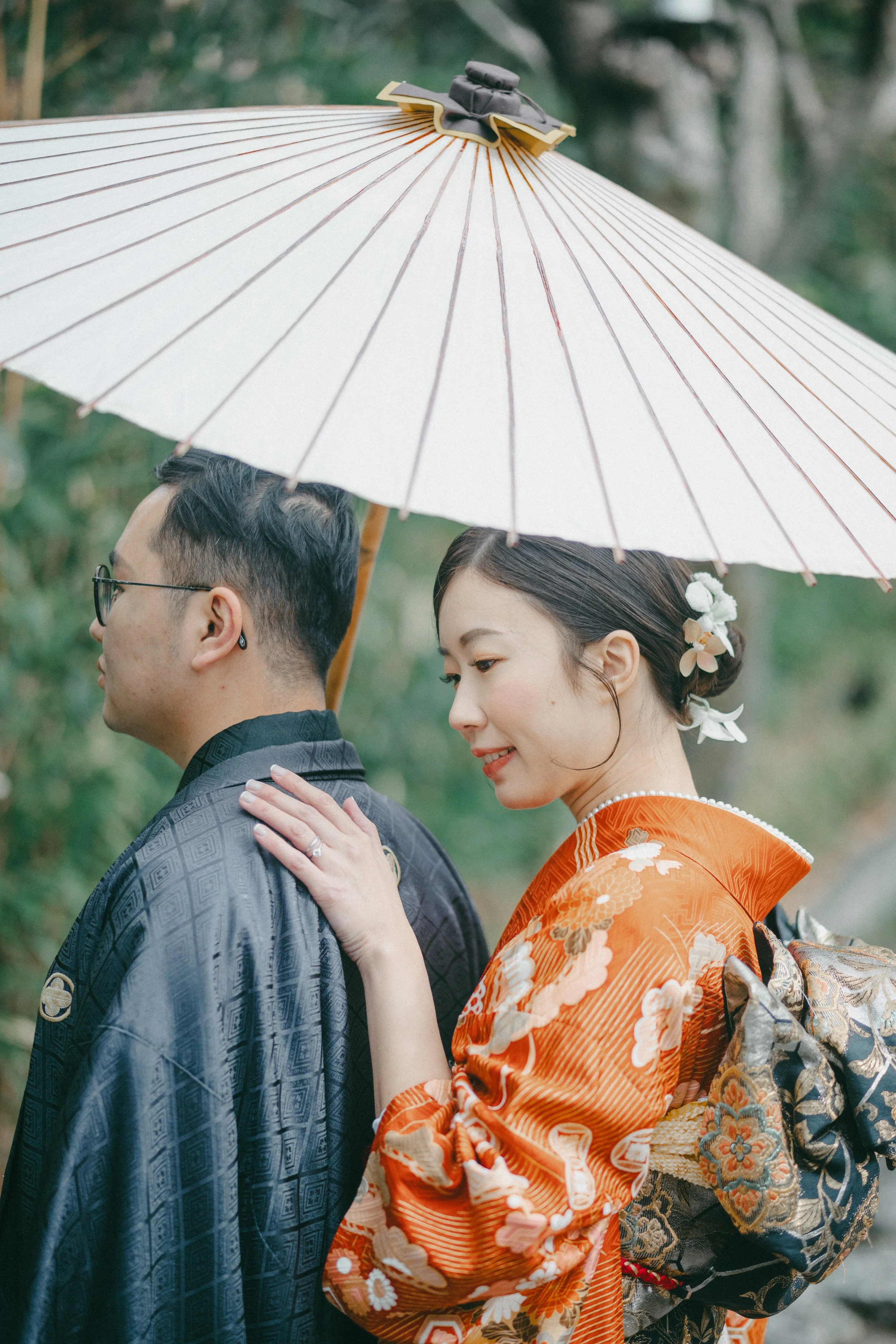 A woman in a traditional Japanese kimono and a man in a dark kimono share an umbrella outdoors, with the woman gently touching the man's shoulder.