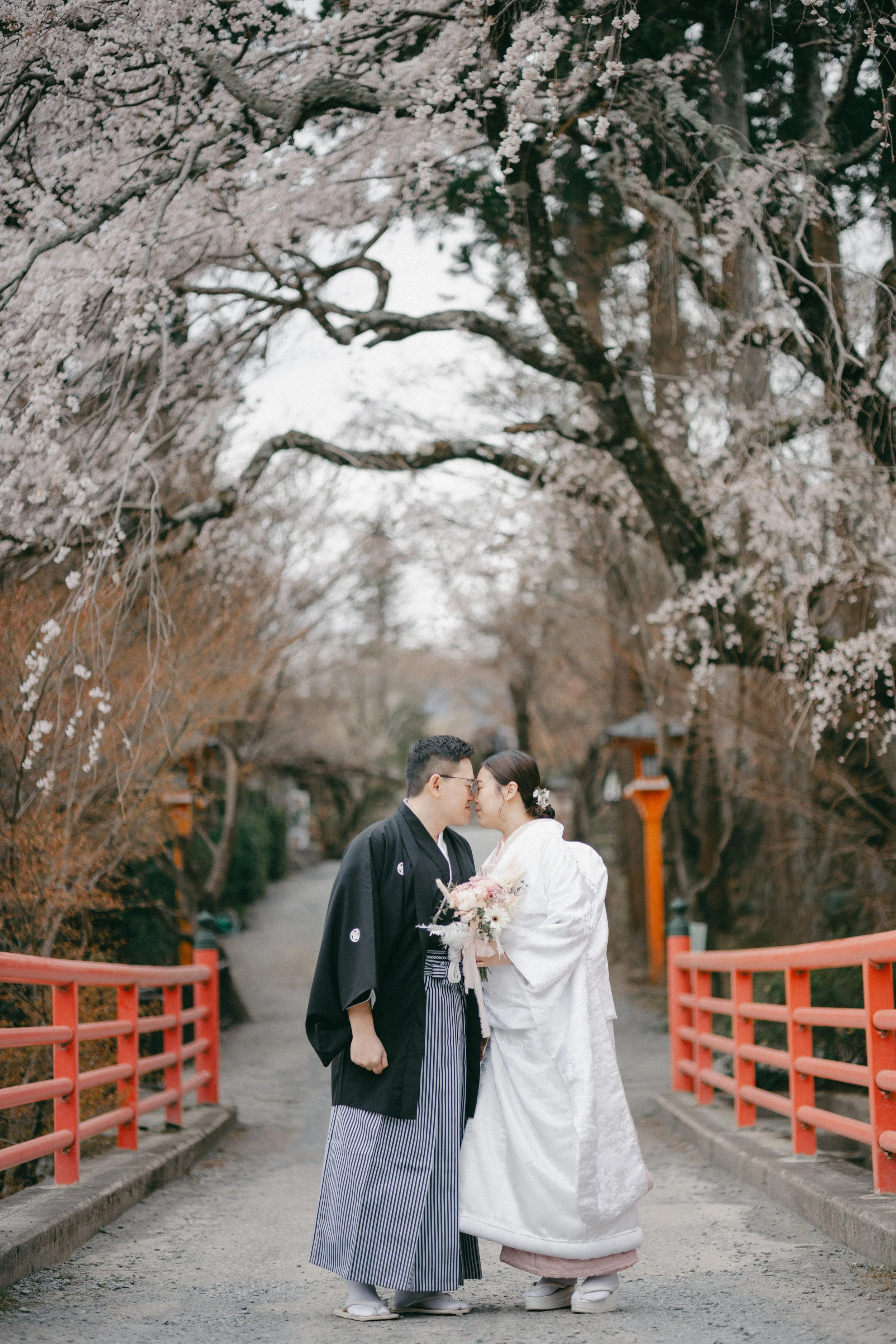 A couple in traditional Japanese wedding attire standing close together on a pathway with cherry blossom trees overhead, holding a bouquet, in a scenic outdoor setting.