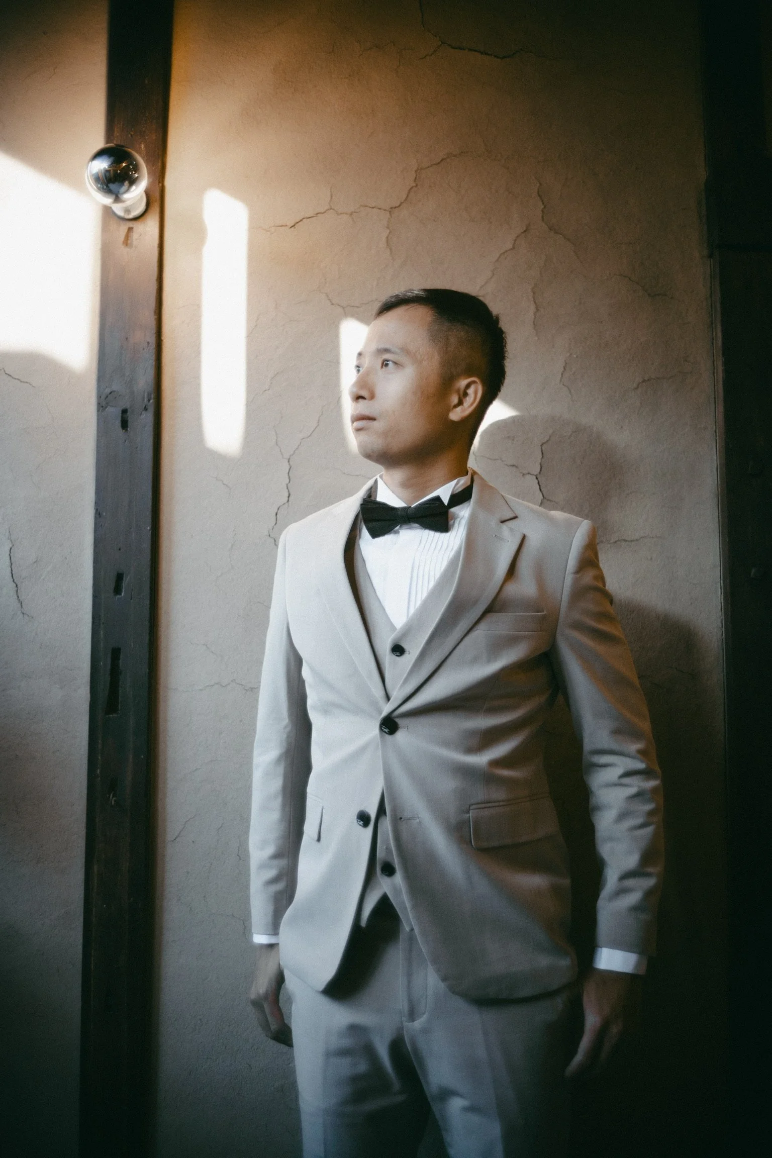 A man in a light-colored tuxedo with a black bow tie standing against a cracked wall, looking to the side with sunlight casting shadow patterns on the wall.