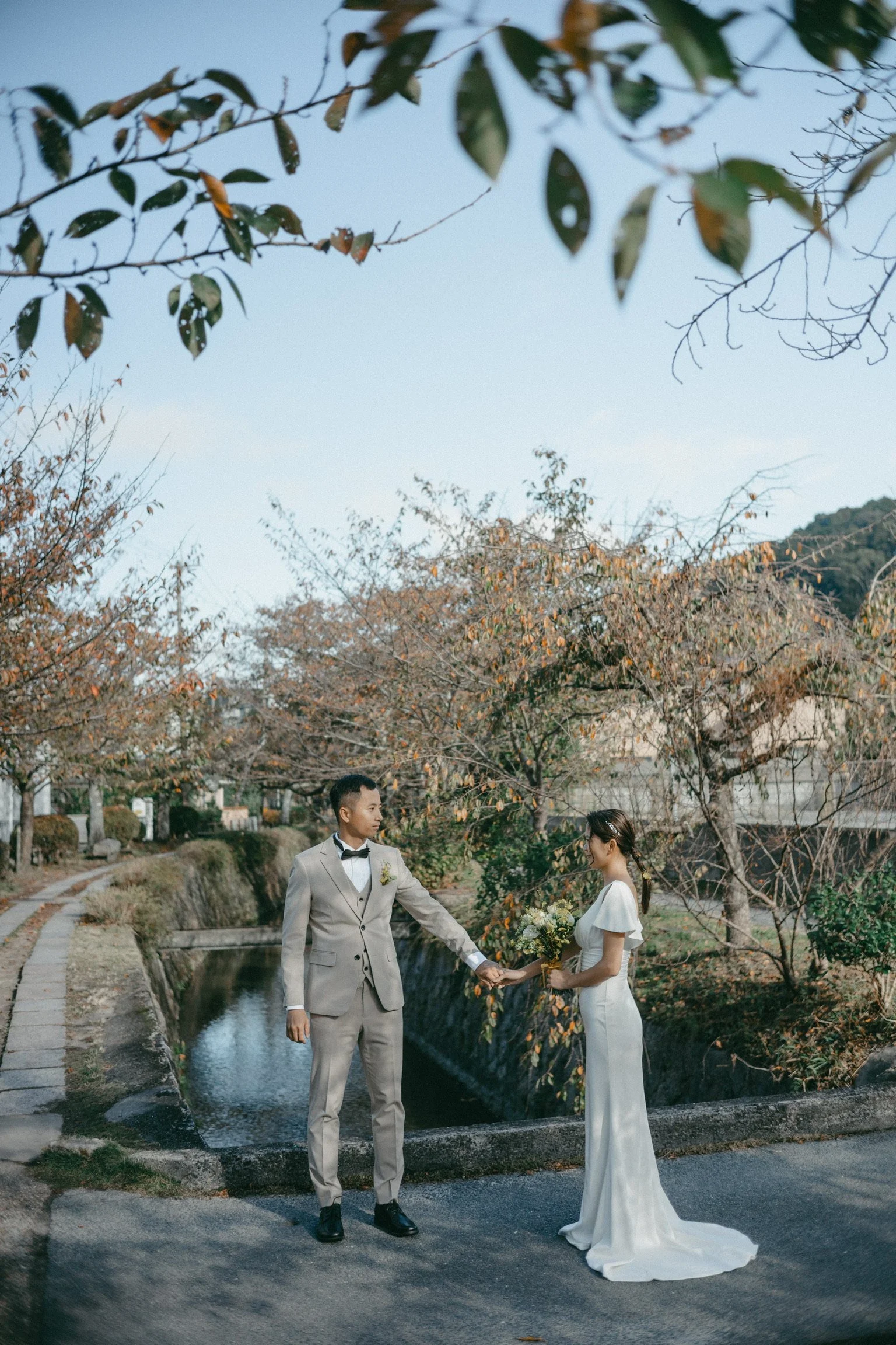 A bride and groom holding hands in an outdoor setting surrounded by autumn trees, with the bride holding a bouquet of flowers and both dressed in wedding attire.