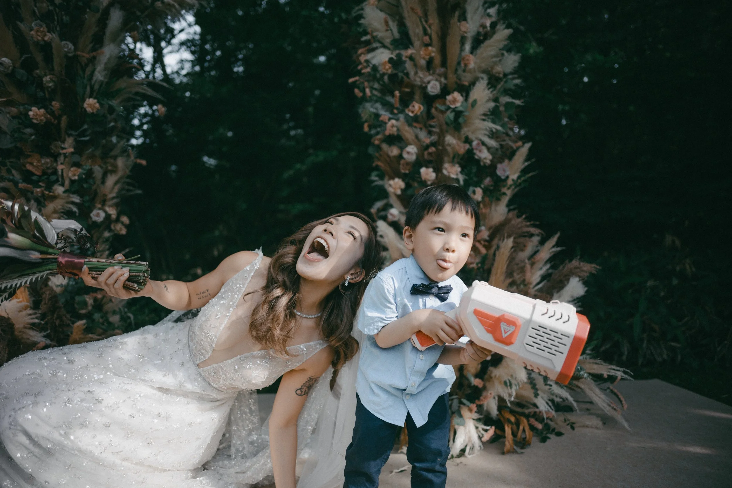 A woman in a wedding dress and a young boy with a toy gun making playful faces outdoors in front of a floral arch.