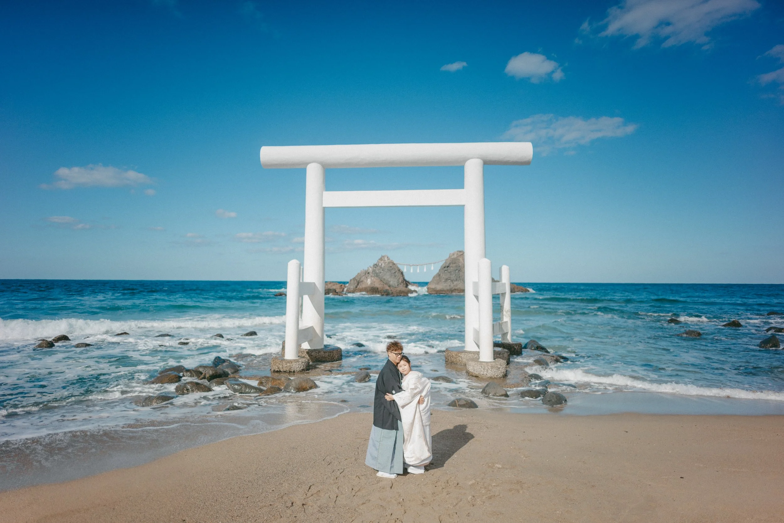 A couple standing on a sandy beach in traditional Japanese wedding attire, with a large white torii gate and rocky islands in the background over the ocean.