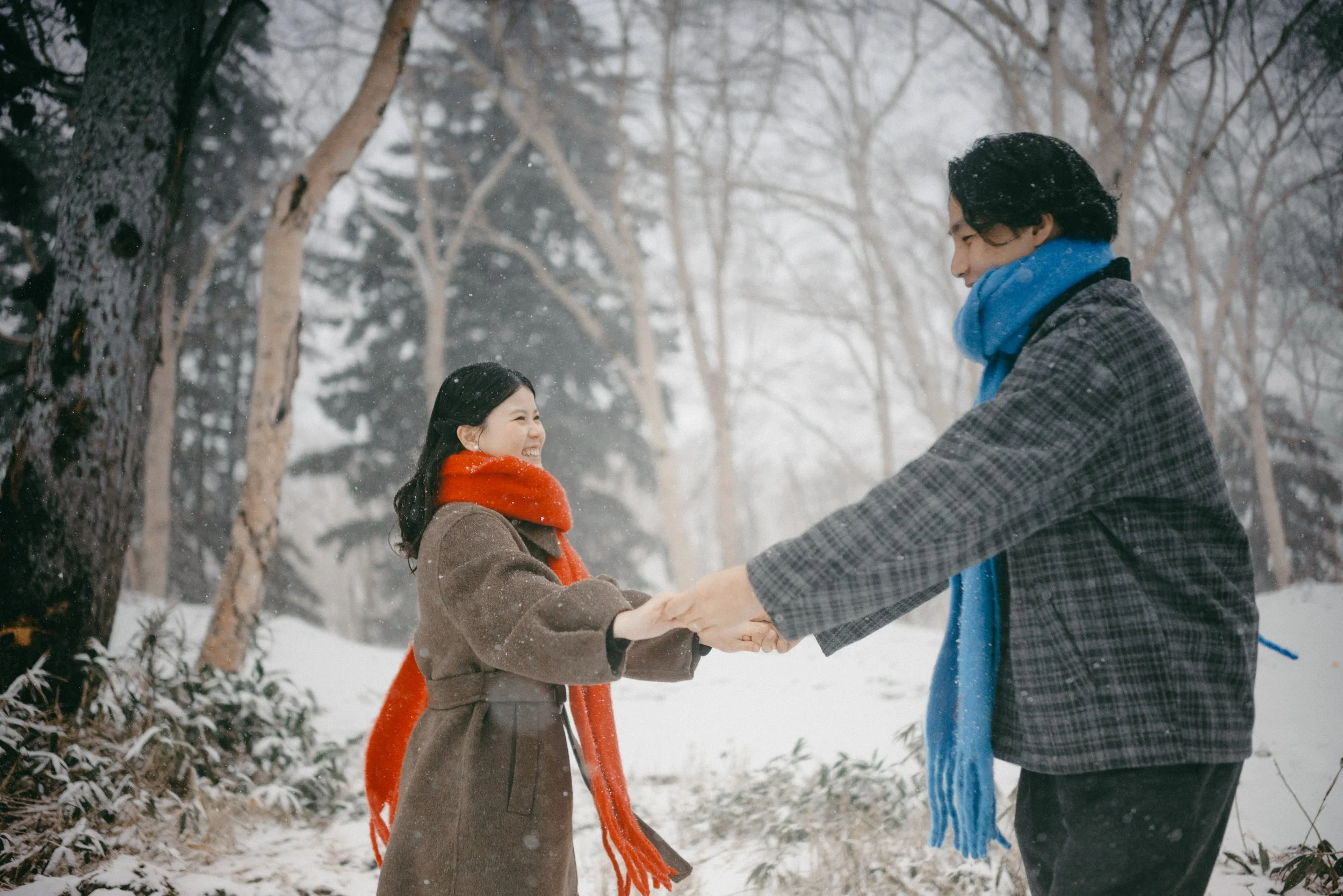A man and woman holding hands and smiling in a snowy forest, dressed in winter clothing.