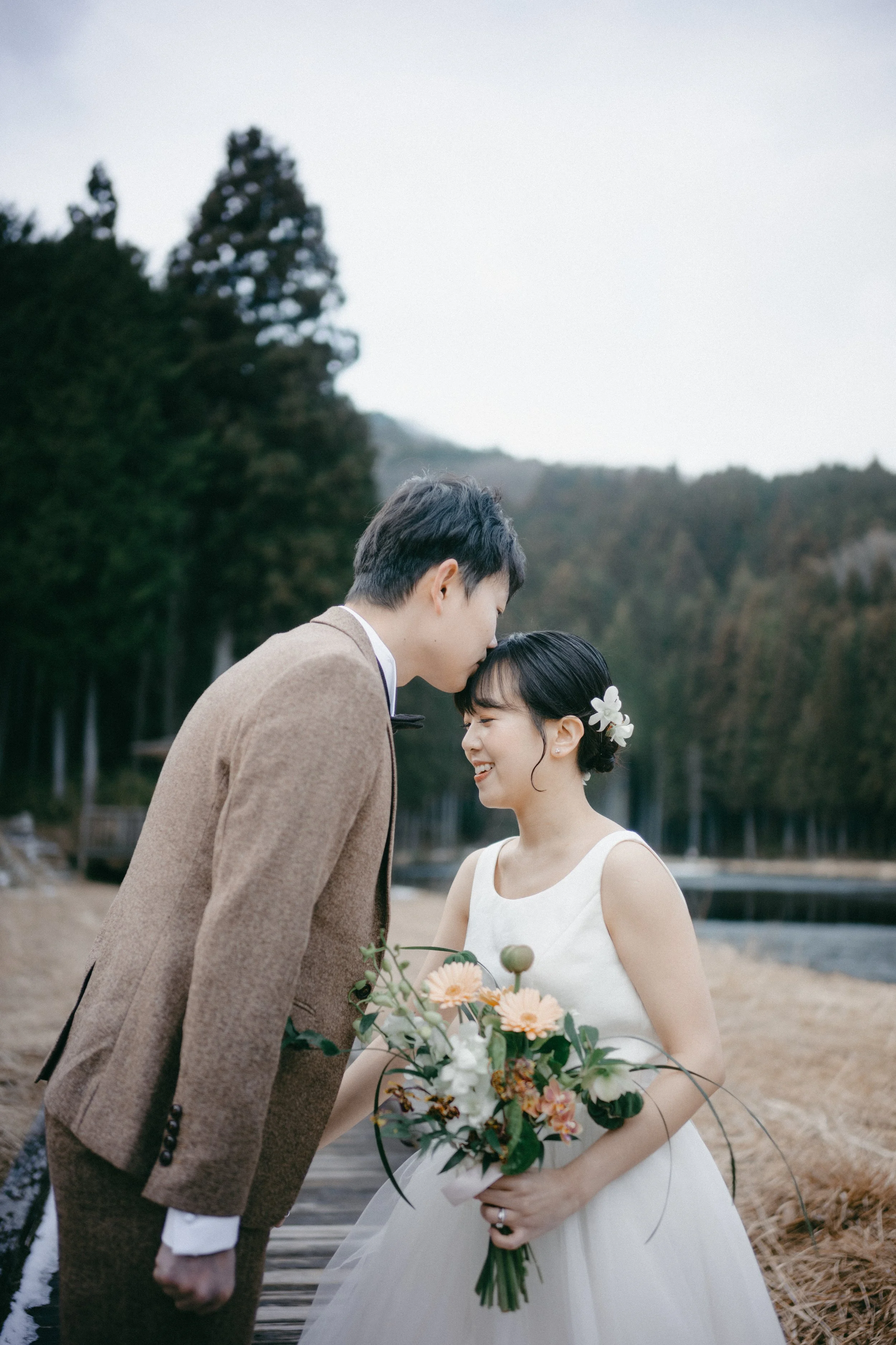 A couple in wedding attire standing on a wooden path outdoors, with the groom kissing the bride on her forehead, the bride holding a bouquet, and smiling.