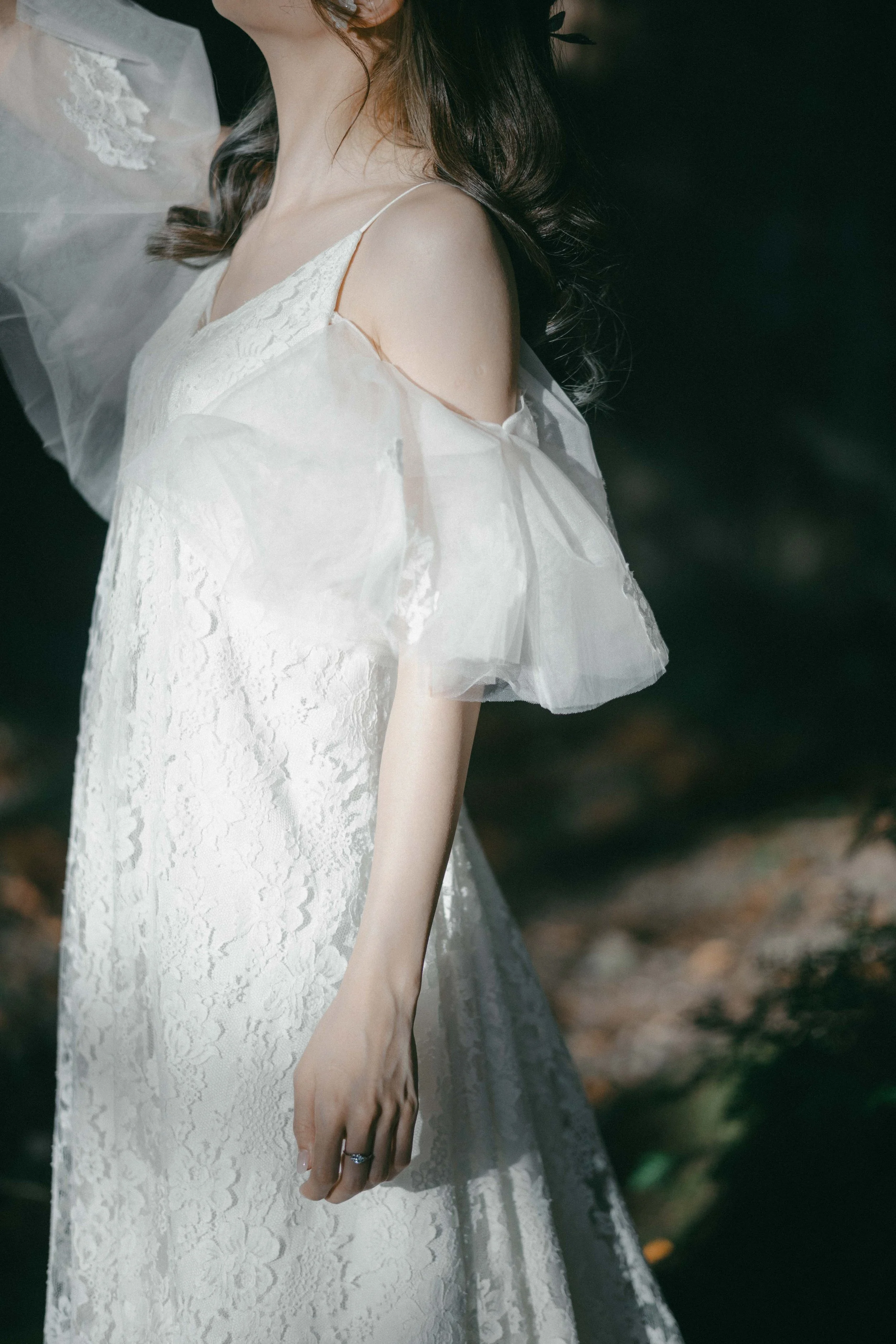 A woman in a white lace dress with off-the-shoulder sheer ruffled sleeves, standing outdoors with dark greenery in the background.